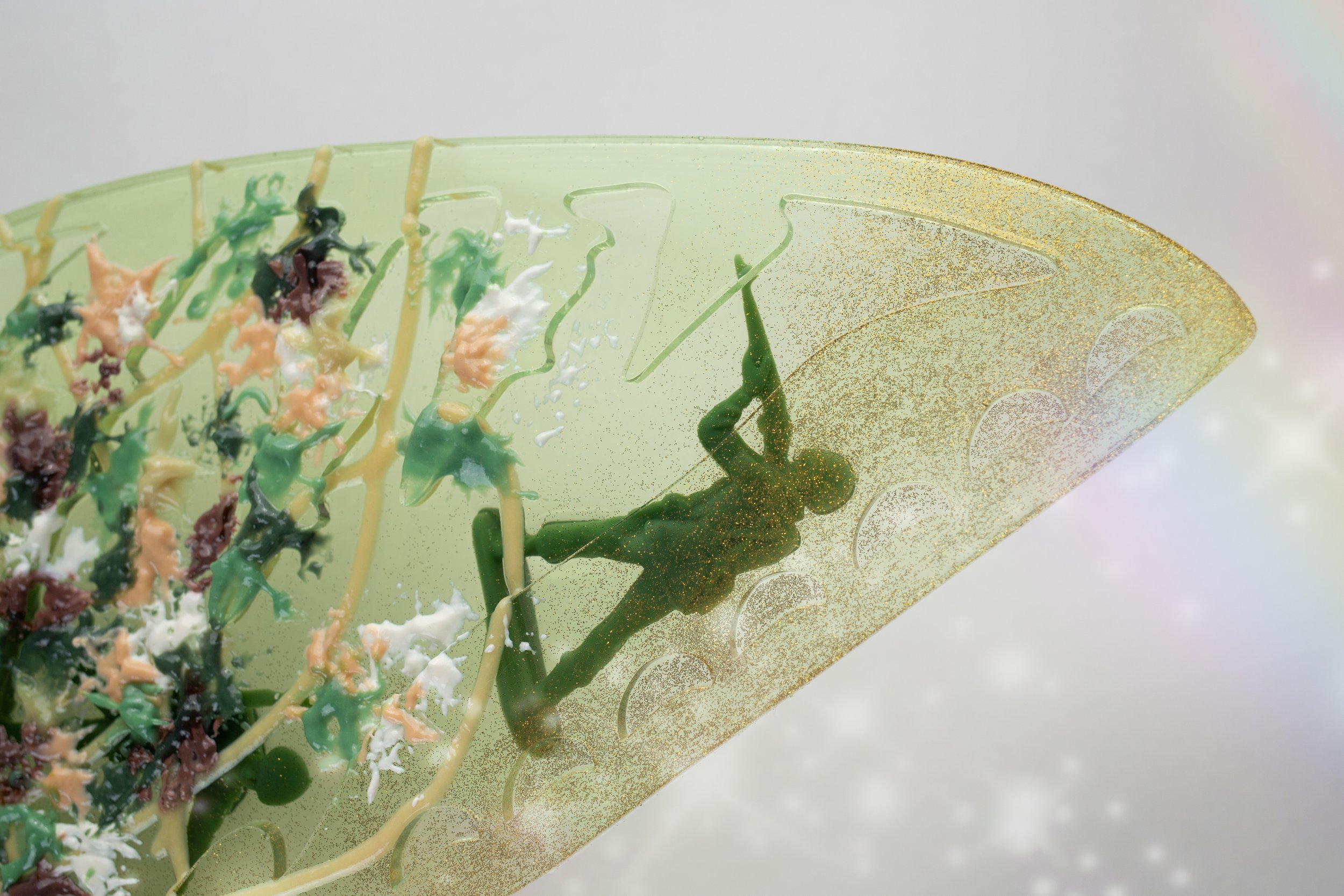 Close-up of a green resin butterfly sculpture with colorful paint splatters and streaks resembling camouflage pattern and an embedded toy army man.