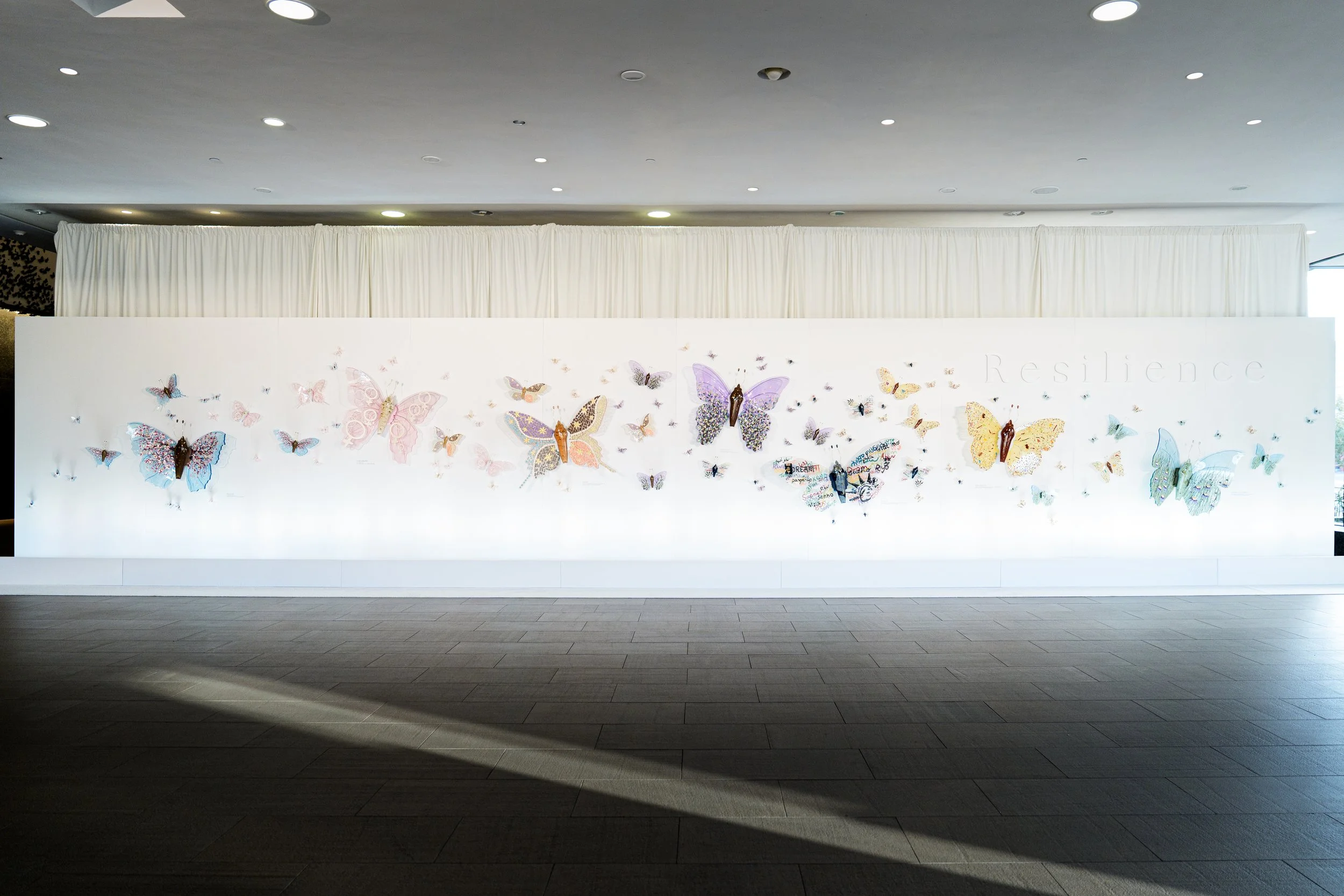 White wall exhibit with colorful resin butterfly sculptures and the word 'Resilience' faintly visible on the right side.