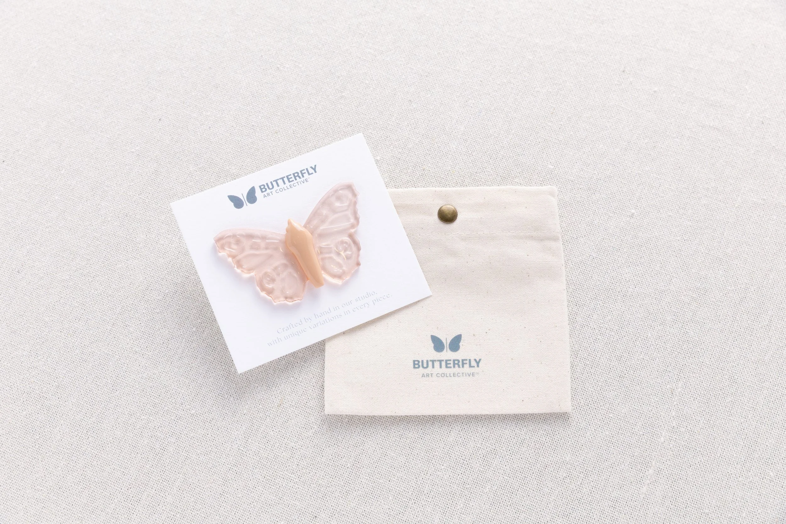 Desert Glass soft resin butterfly pin in translucent blush peach with a warm peach body, displayed on a branded card with a cotton canvas pouch.