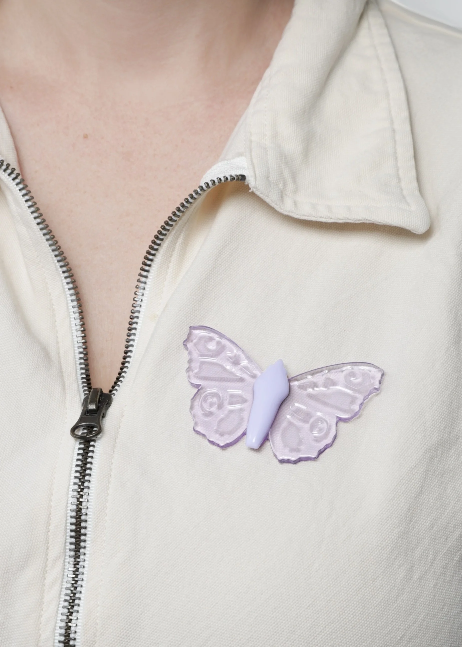 Hand-cast lavender soft resin butterfly pin attached to a cream canvas jumpsuit, featuring translucent, glass-like wings and a sculptural design by Butterfly Art Collective.
