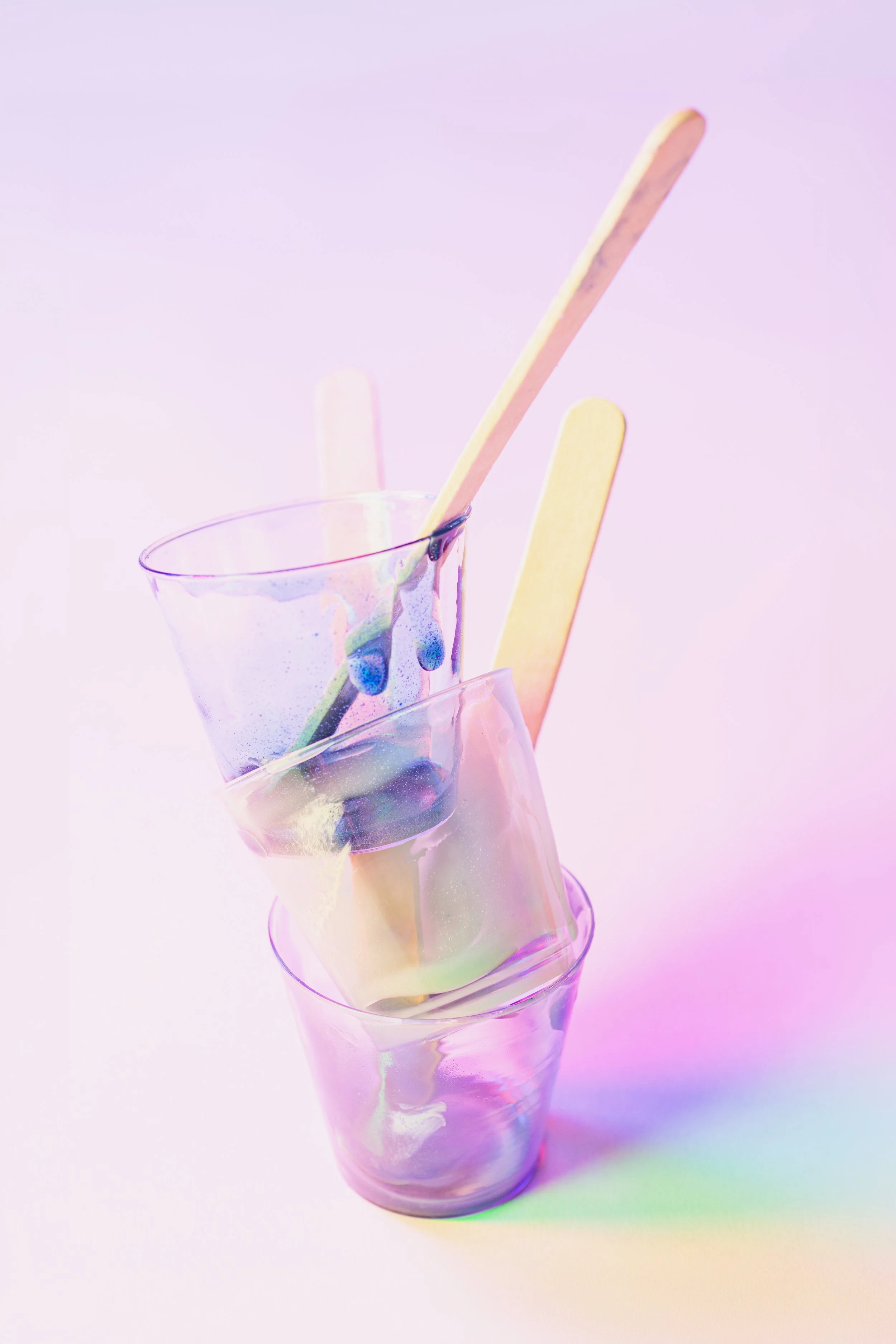 Stacked transparent glasses with colorful resin splatters and three pastel-colored popsicle sticks on a pink and purple gradient background.