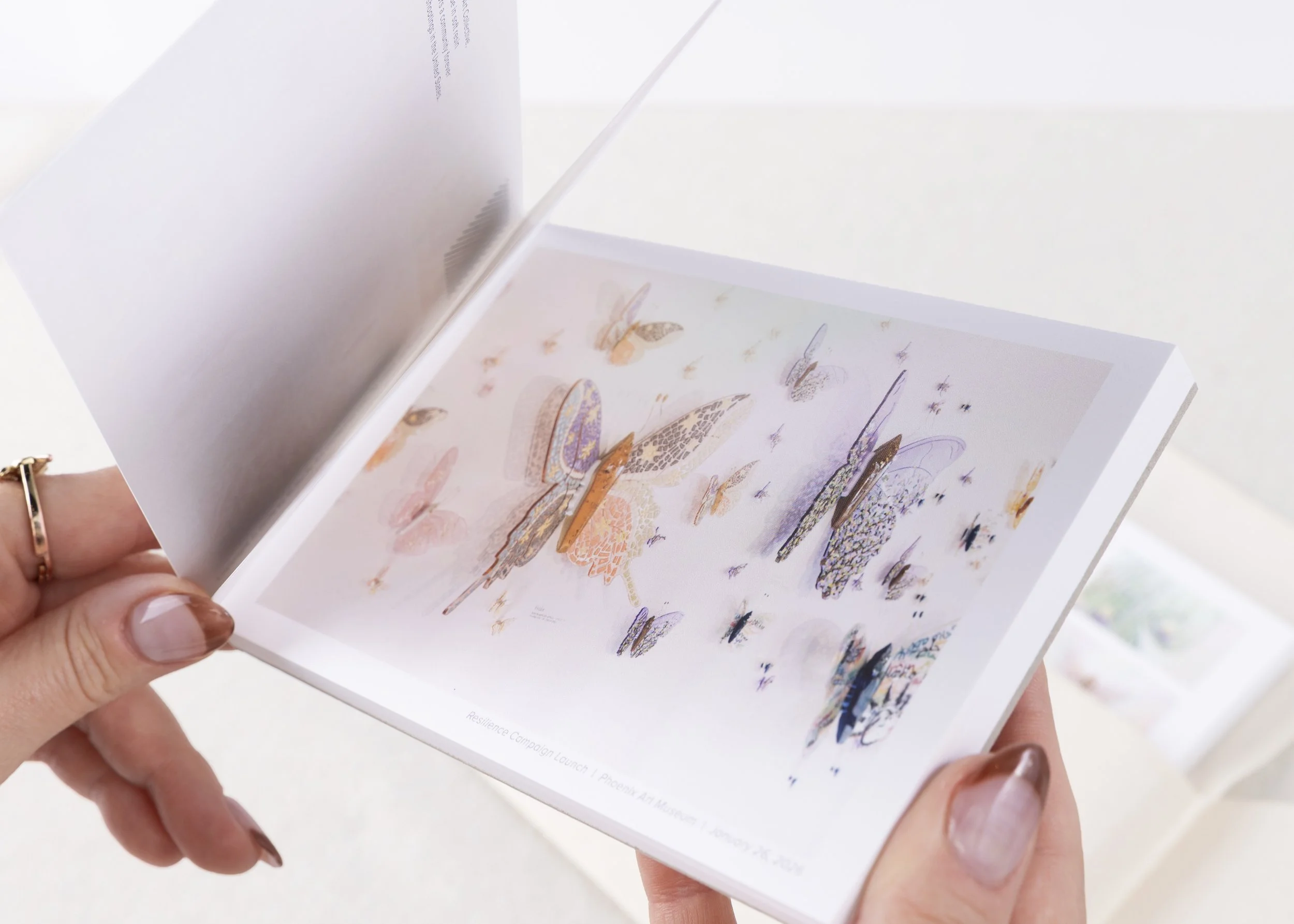 Hands flipping through the Resilience Postcard Book featuring colorful butterfly artwork inspired by the Butterfly Art Collective's sculptural collection.