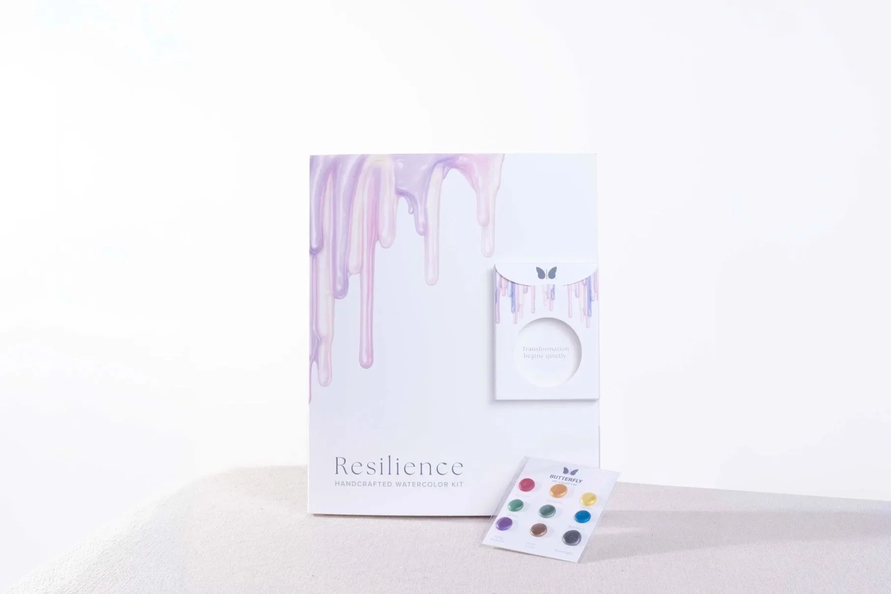 Resilience watercolor kit packaging featuring soft lavender paint drips, a metallic watercolor palette card, and a small circular pocket displayed together.