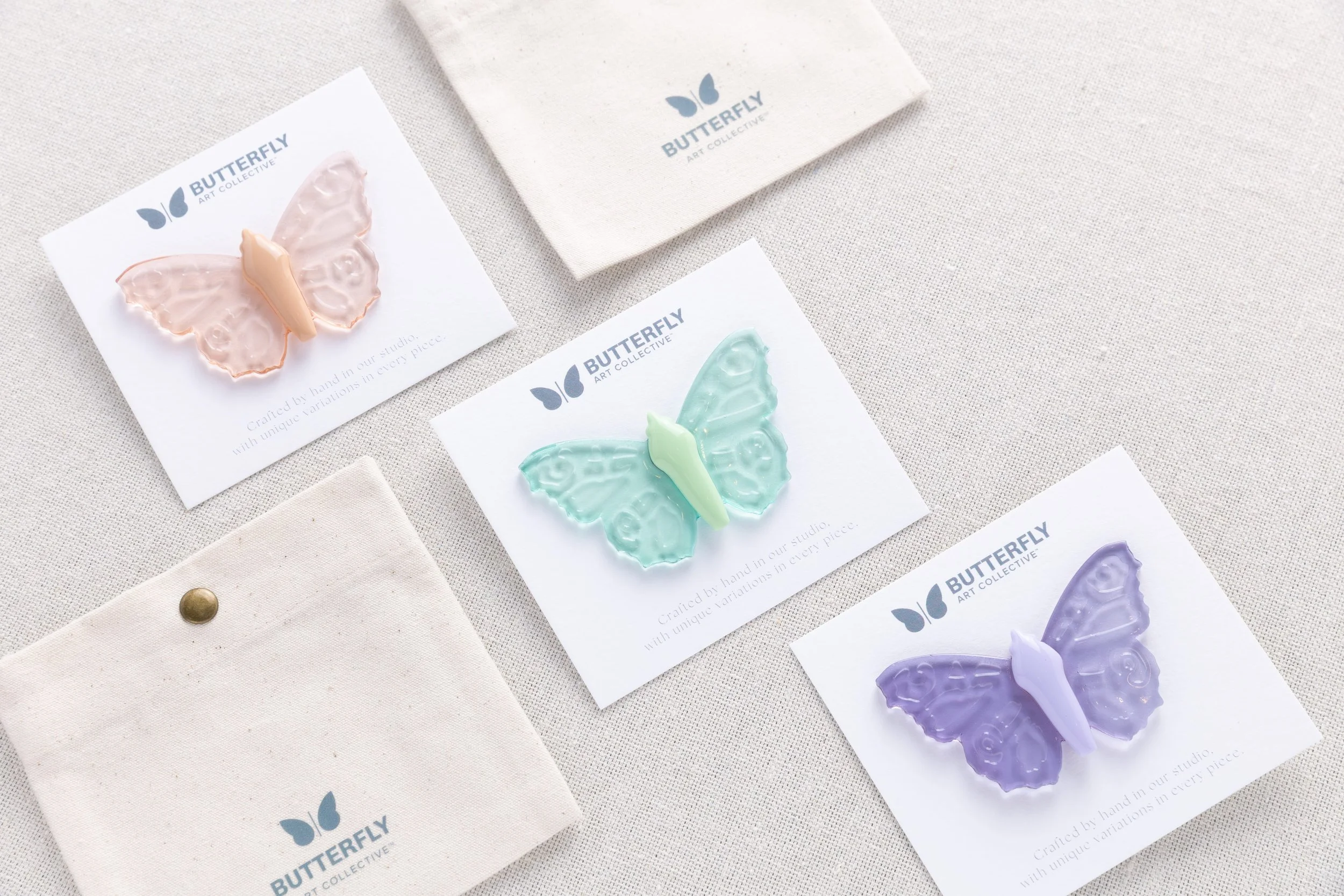 Hand-cast soft resin butterfly pins by Butterfly Art Collective in Twilight Glass, Sea Glass, and Desert Glass, displayed with branded cotton canvas pouches.