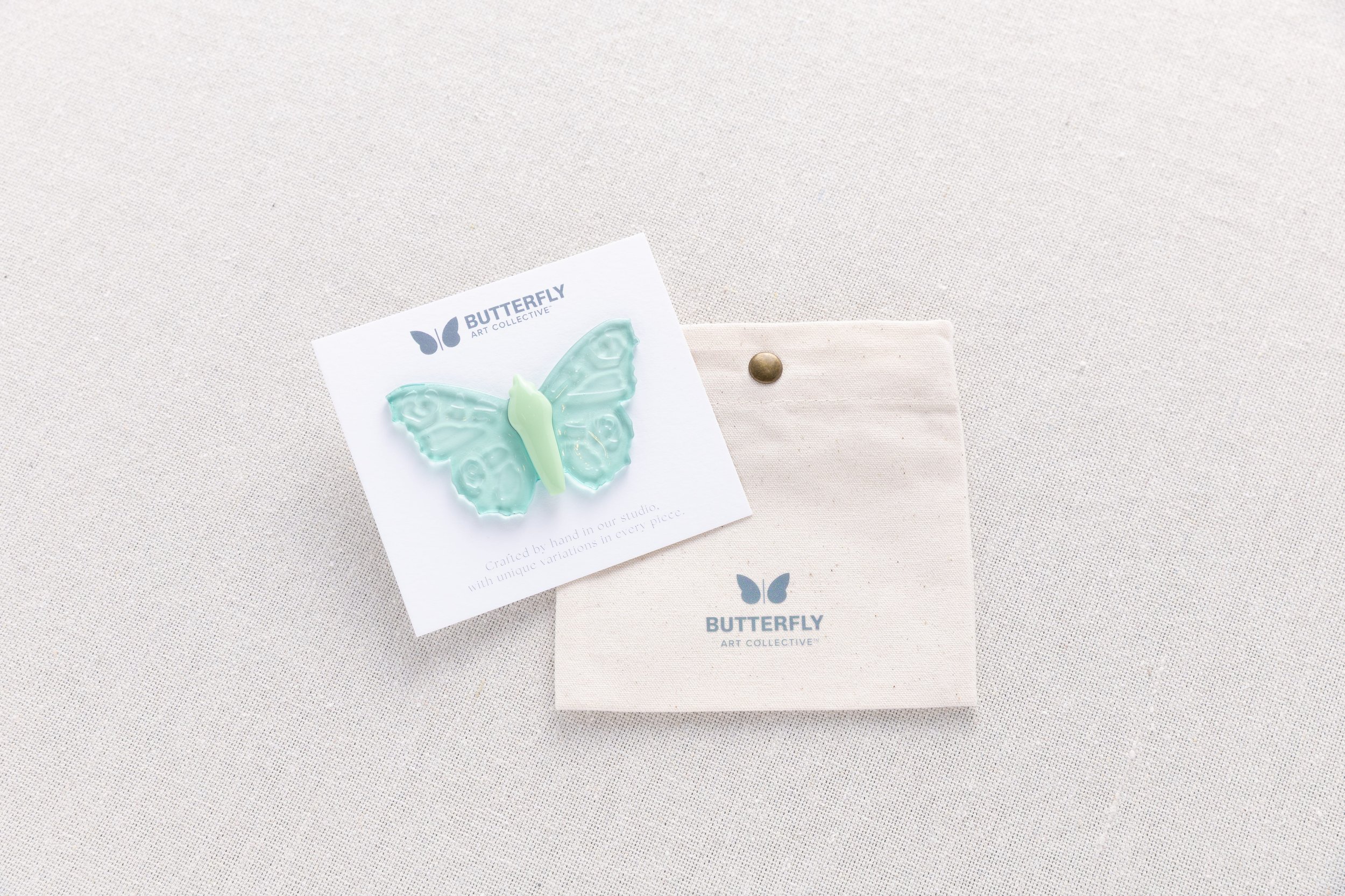 Sea Glass soft resin butterfly pin in translucent aqua with a soft mint-green body, displayed with Butterfly Art Collective's cotton canvas pouch.