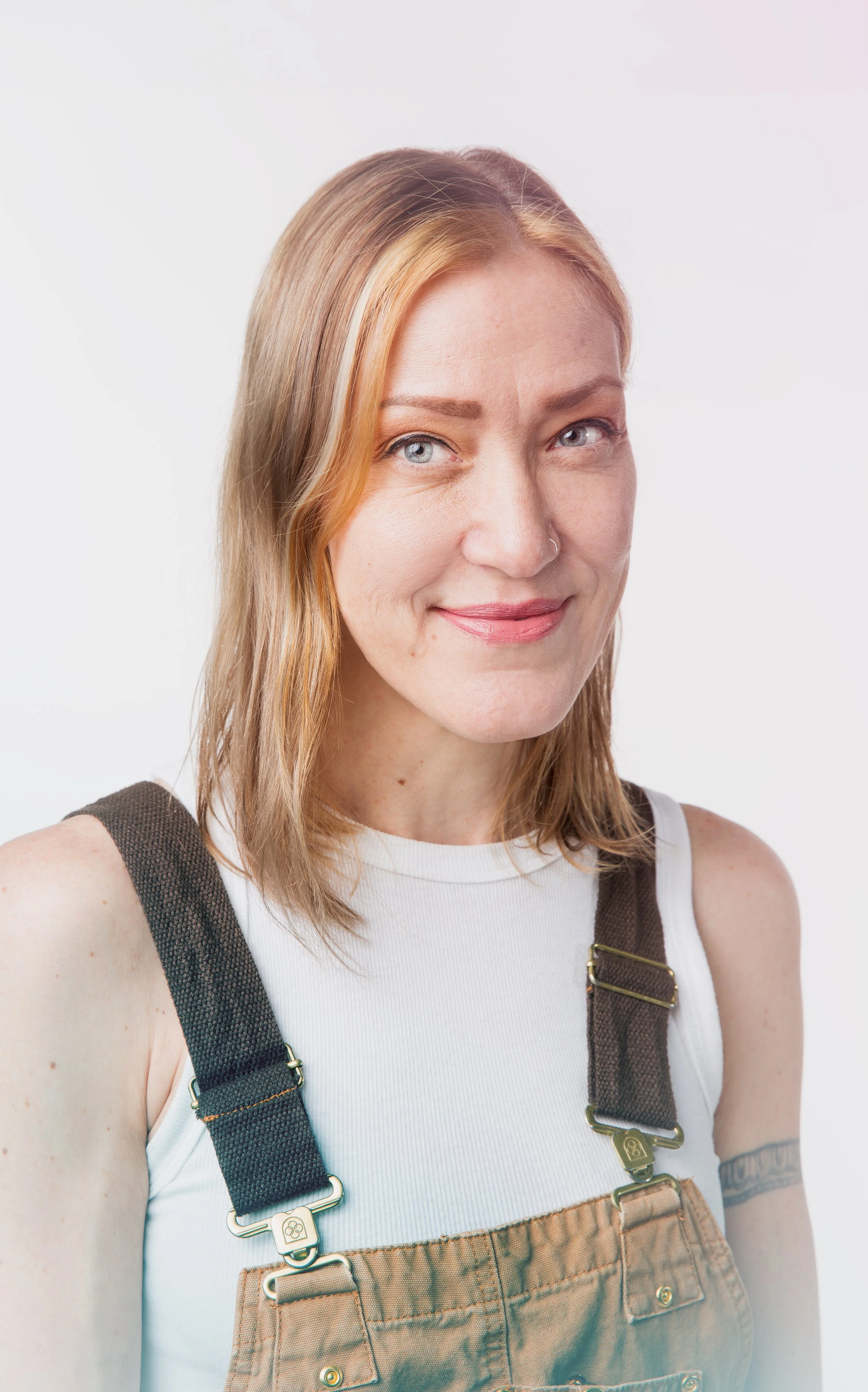 Portrait of Brandi Read with shoulder-length blonde hair, blue eyes, and a nose piercing, smiling and wearing a white tank top and brown overalls with black straps.