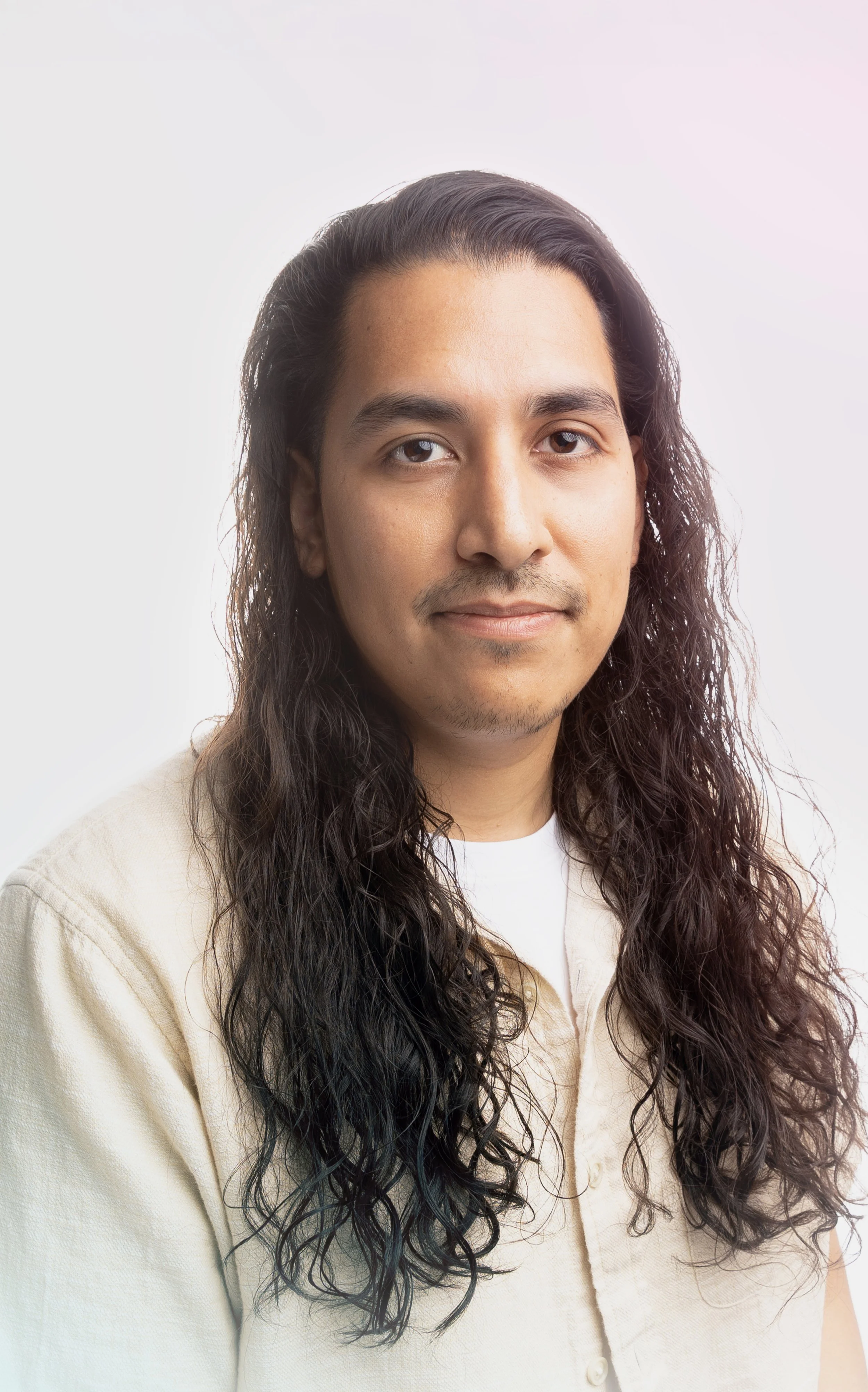 Portrait of Julian Silva with long wavy black hair, wearing a cream-colored jacket and white shirt, against a plain light background.