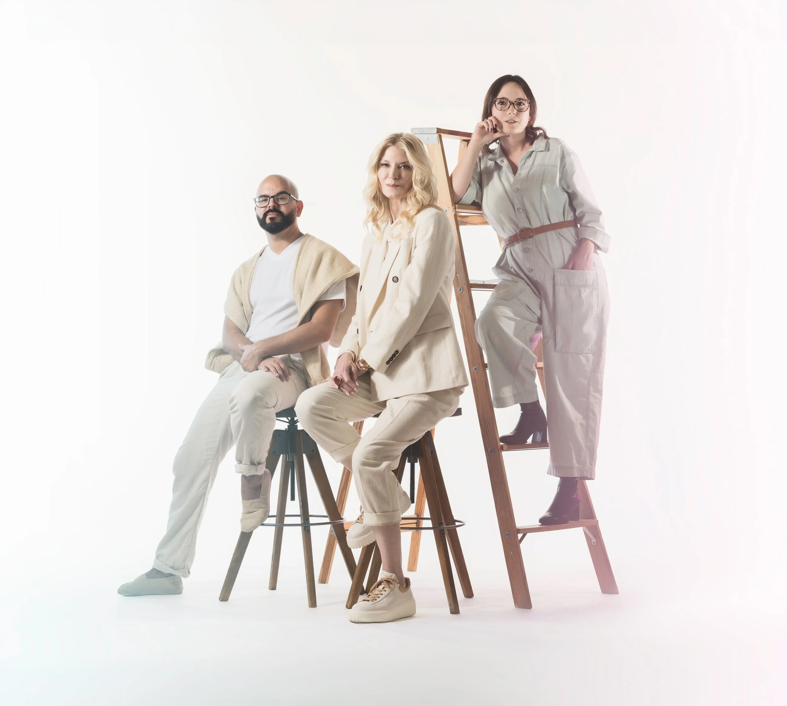 Three artists posing together against a white background. Artist Cheryl Najafi is sitting in the center on a stool with a ladder behind her, Kelsey Vinas is standing on the ladder, and Gabriel Vinas is sitting on a chair with his arms crossed.
