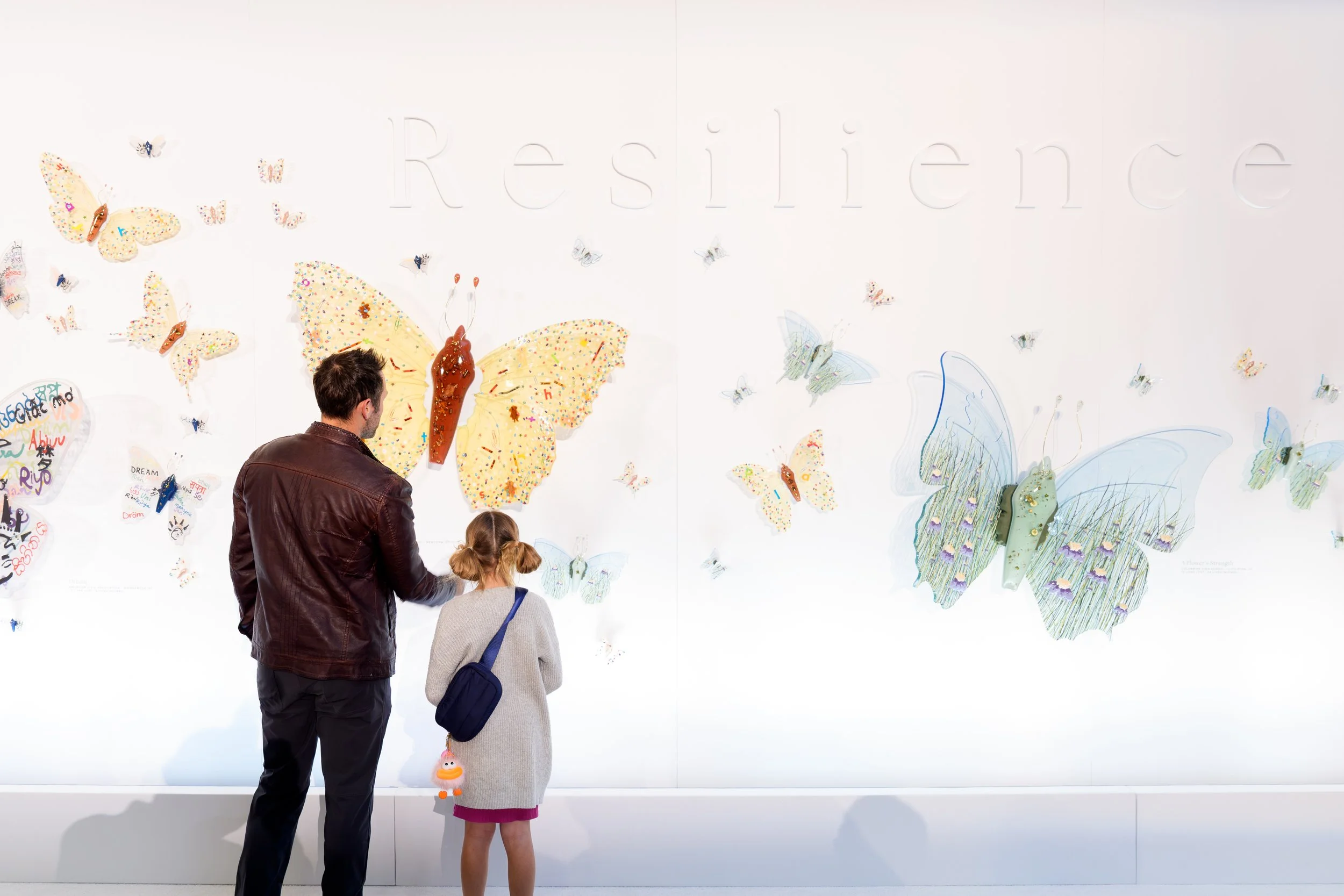 A man and a young girl standing in front of a white wall with large, colorful resin butterfly sculptures. The wall has the word "Resilience" embossed on it.