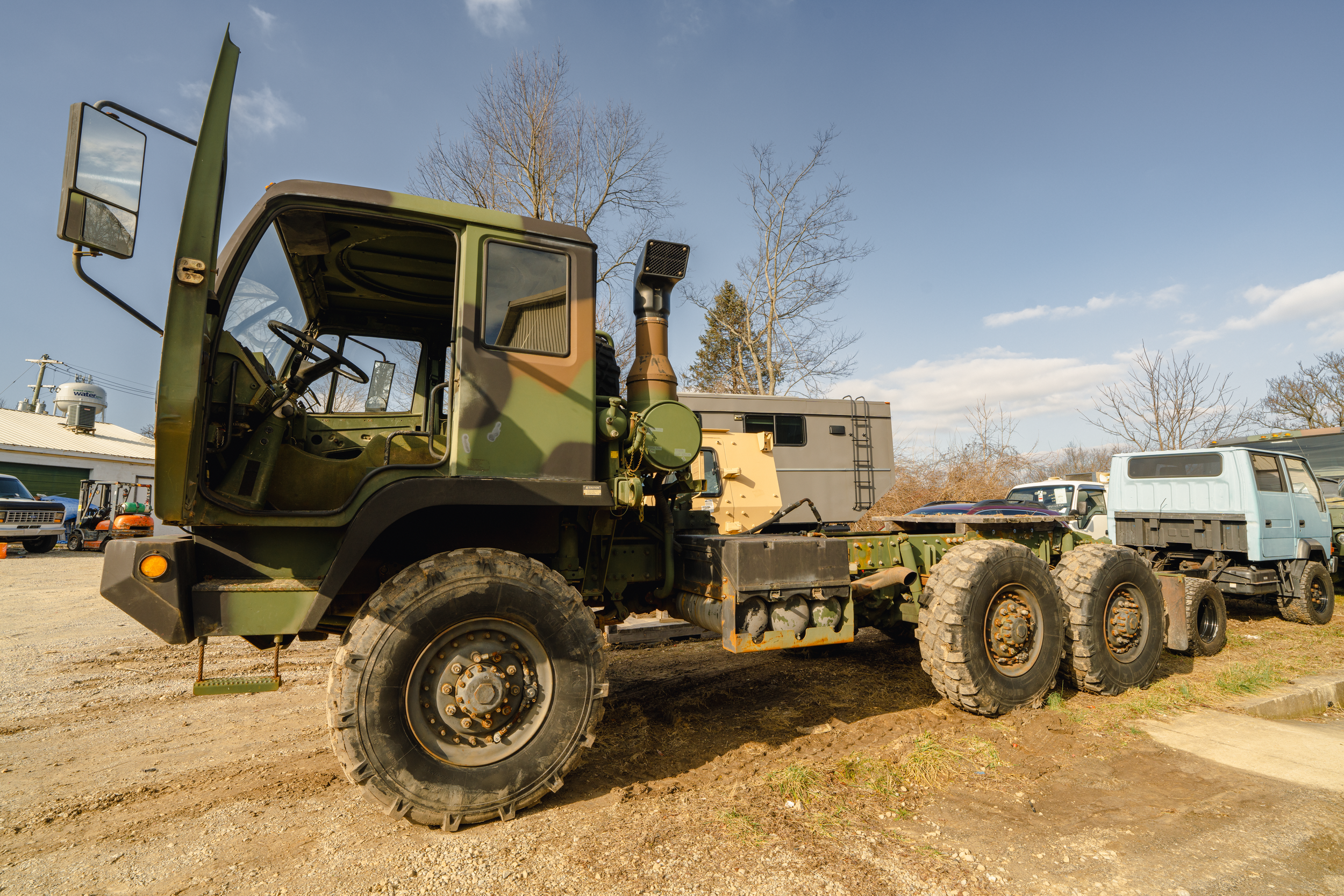 A large military-style truck with a camouflage pattern parked on a dirt lot, with other vehicles and leafless trees in the background under a partly cloudy sky.