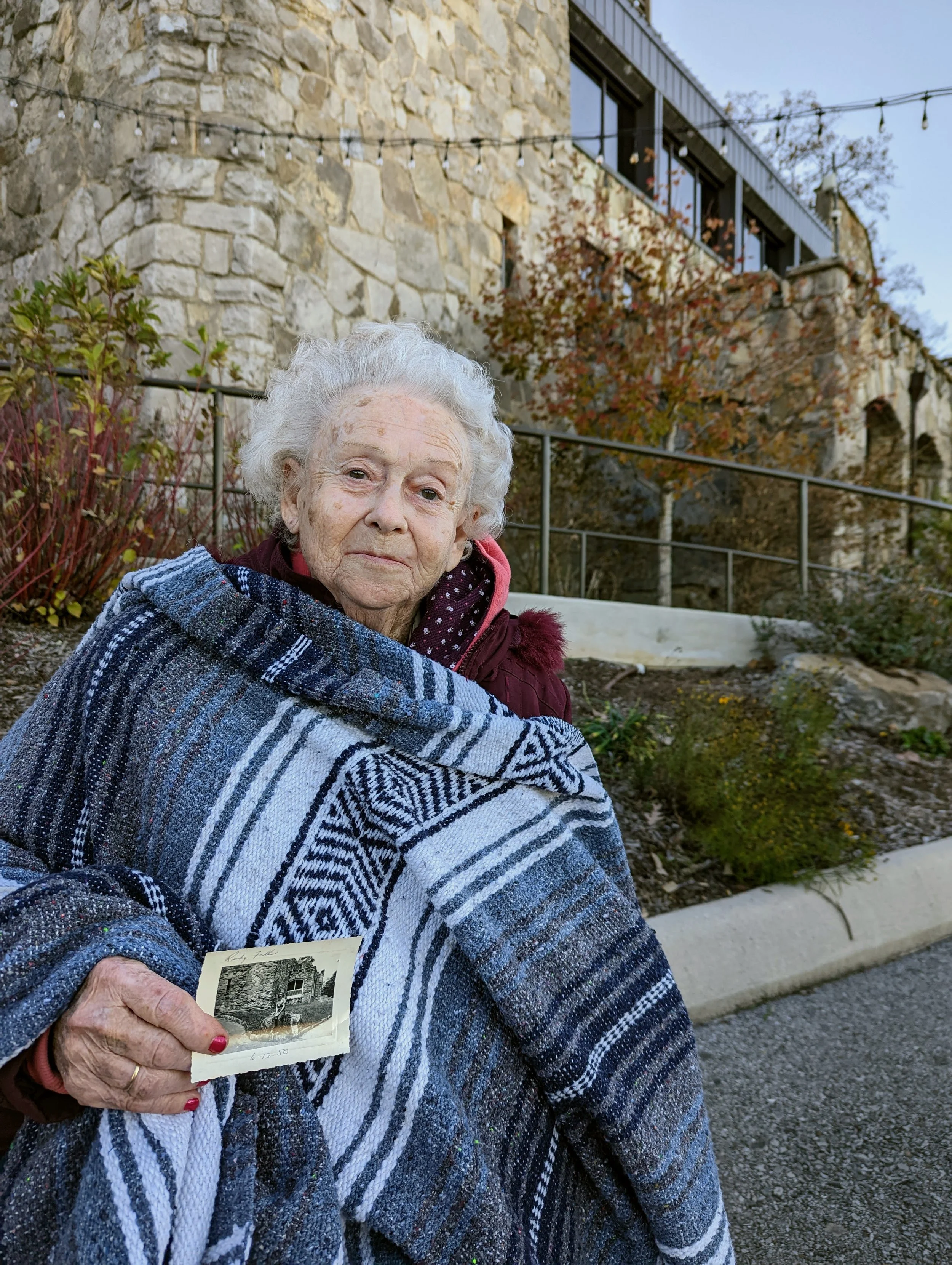 An elderly woman with grey curly hair, wearing a maroon jacket, is holding a black and white photograph while wrapped in a striped woven blanket outdoors on a cloudy day. Behind her is a stone building and a garden with autumn foliage.