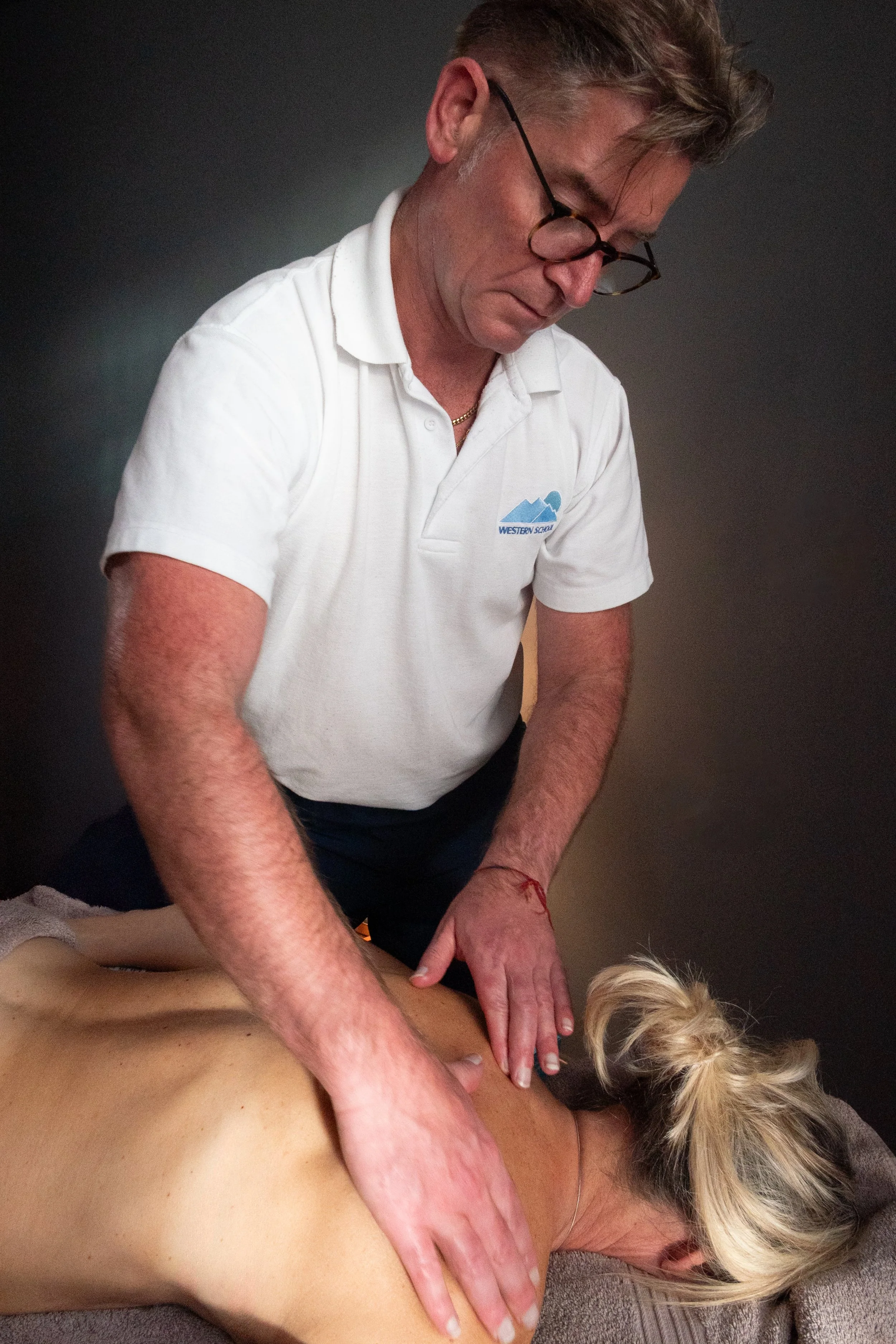 A man wearing glasses and a white polo shirt giving a massage to a woman lying face down on a massage table.