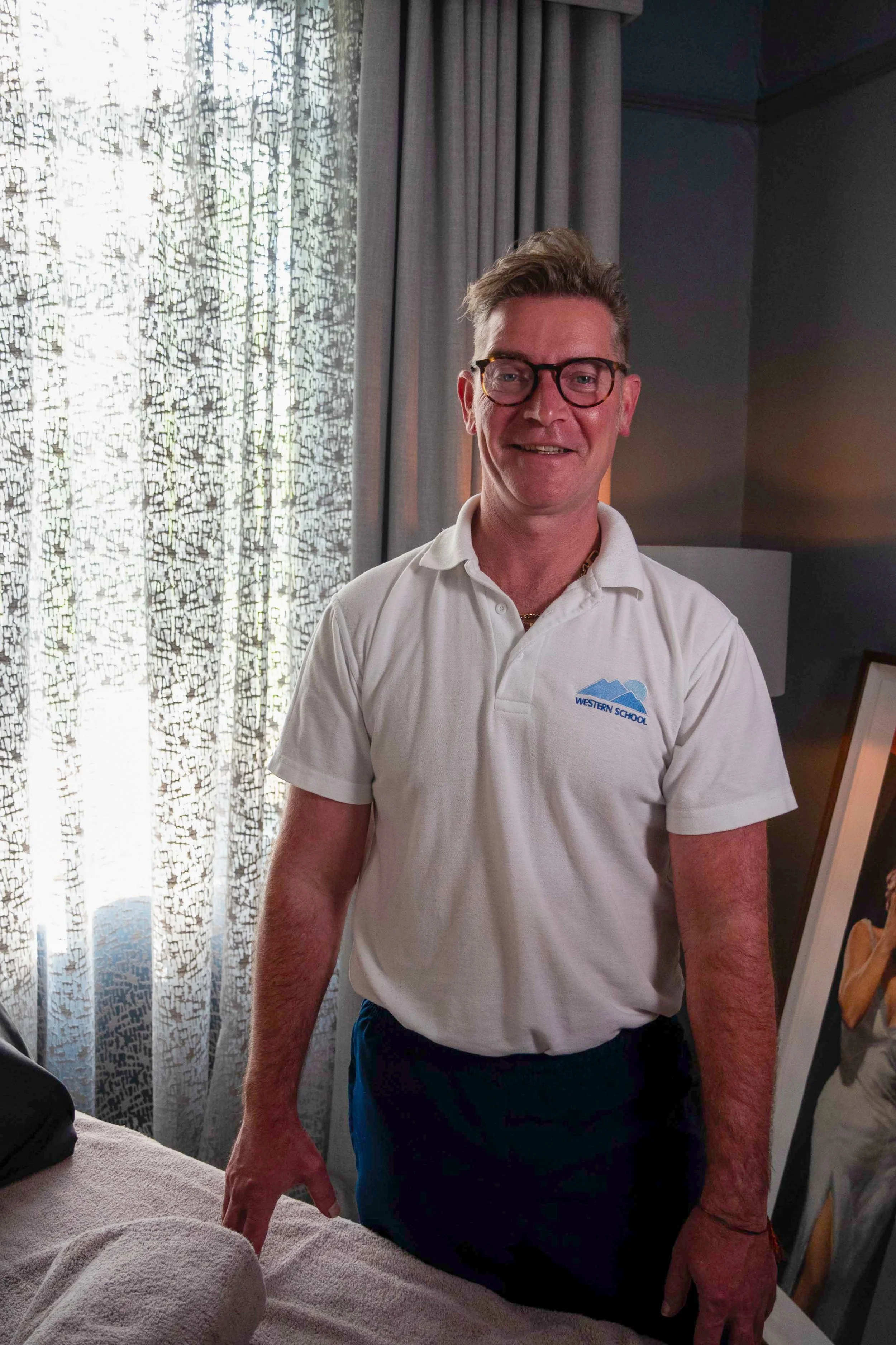 A man with glasses, short blond hair, and a friendly expression is standing in a room near a window with patterned curtains. He is wearing a white polo shirt with a 'Western School' logo and dark pants.