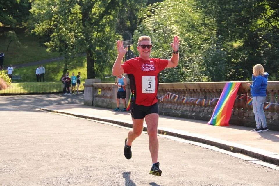 A man running a race, wearing a red shirt, black shorts, sunglasses, and a bib number 5 that says Shannon, with trees and a stone fence decorated with rainbow flags in the background.