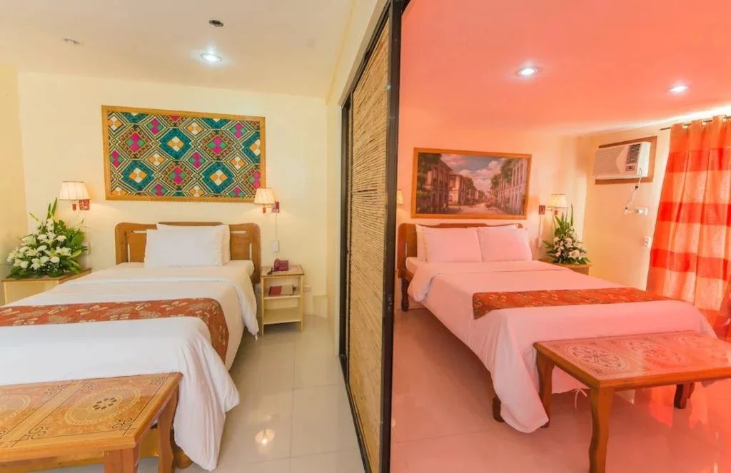 Interior of a hotel room with two separate beds, each with white bedding and decorative throws. The room features colorful wall art, bedside tables with lamps, floral arrangements, curtain, and an air conditioning unit.