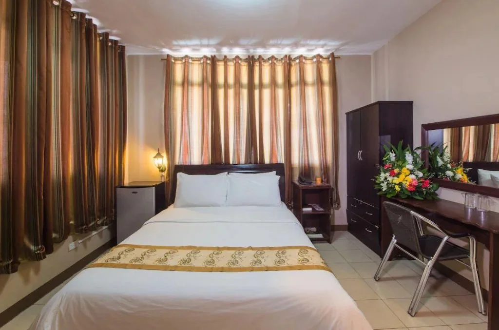 Hotel room with a neatly made bed, curtains, a dresser with a mirror, a chair, a small refrigerator, and a bouquet of flowers on the furniture.