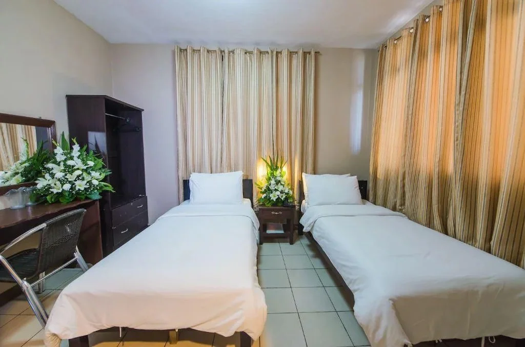 Hotel room with two twin beds, white bedding, wooden nightstand, large floral arrangements, and beige curtains.