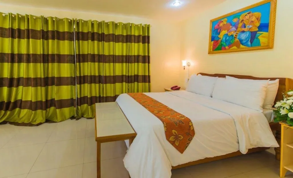A hotel room with a large bed, colorful artwork on the wall, yellow and green striped curtains, and a bedside lamp.