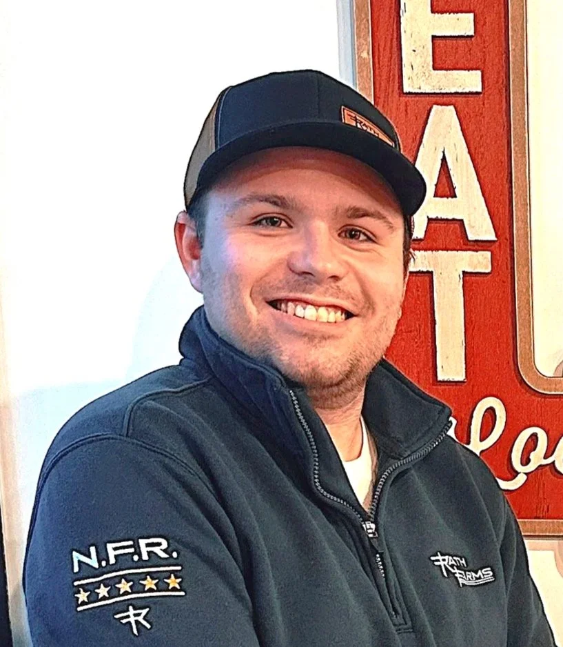 A person sitting at a desk with a computer, wearing a dark hoodie, and smiling. There's a nameplate on the desk.