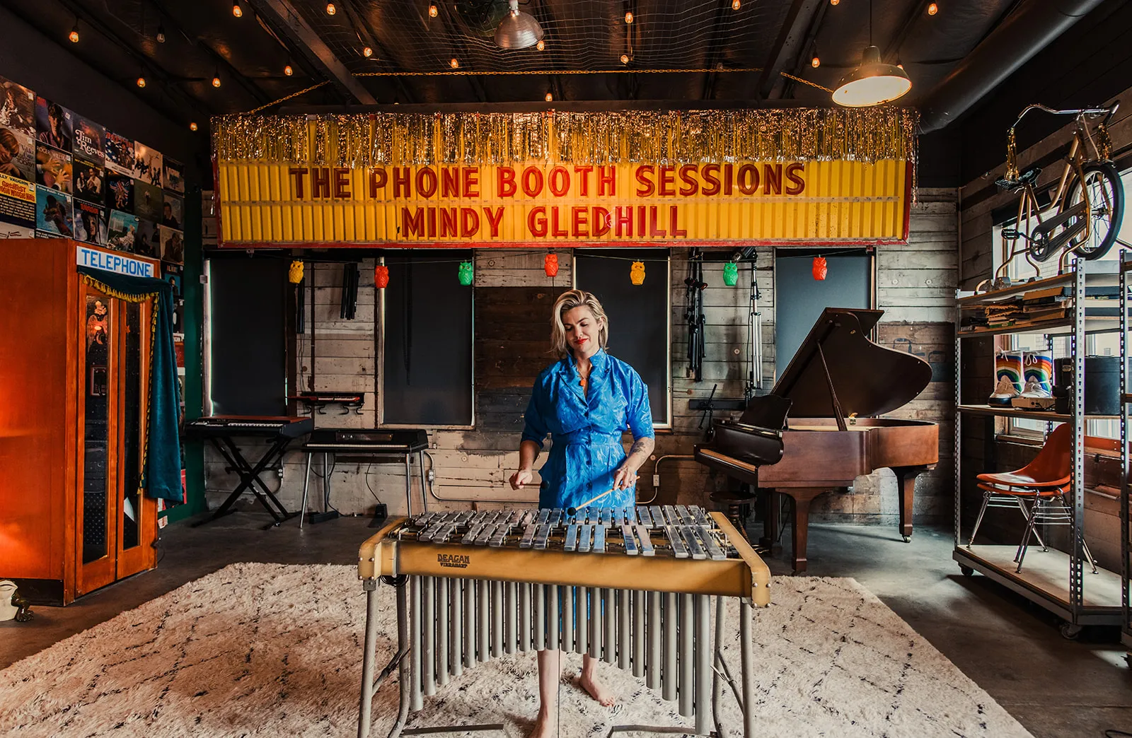 A woman in a blue dress playing a vibraphone in a music studio. A large sign overhead reads 'The Phone Booth Sessions Mindy Gledhill.' The studio has a piano, a bicycle mounted on the wall, shelves with records, and various musical equipment.