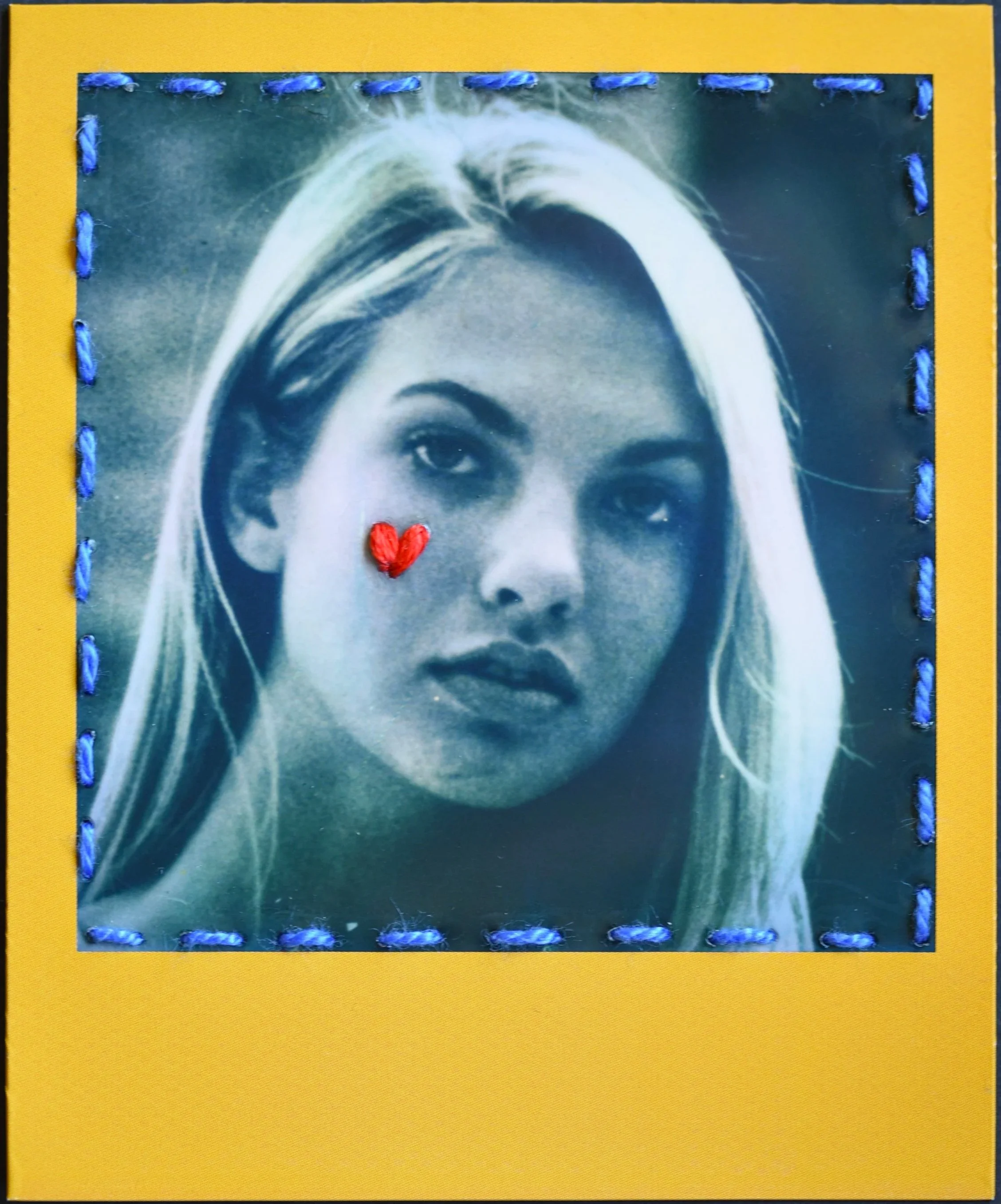 A black and white portrait of Mindy Gledhill with long hair, with a red stitched heart near her cheek, framed with blue stitches on a yellow background.