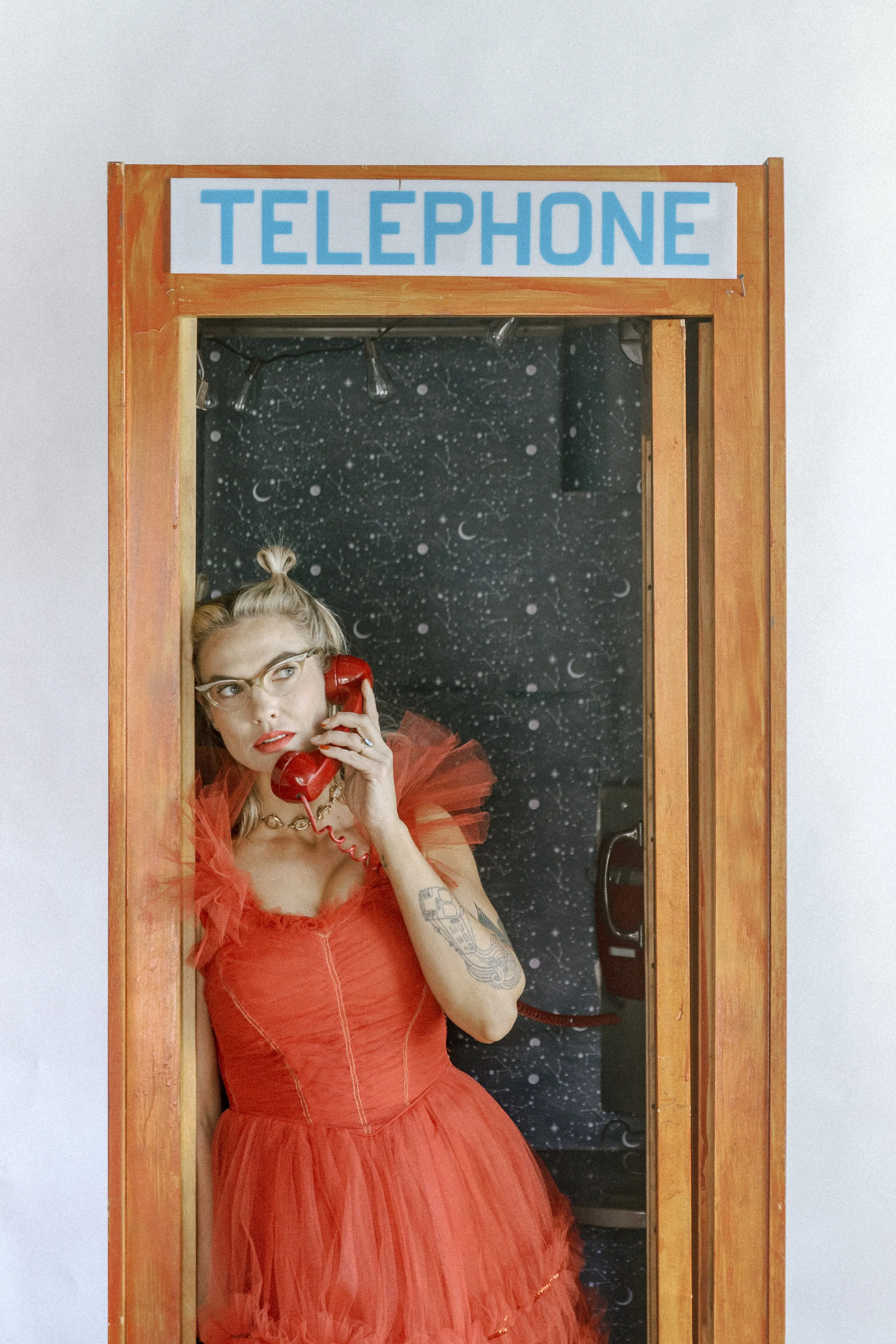 Mindy Gledhill, an indie singer-songwriter, in a red dress with glasses and tattoos talking on a red payphone inside a wooden phone booth with a space-themed background.