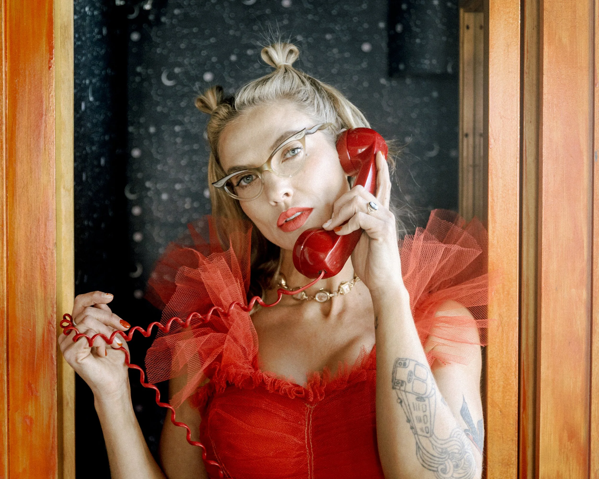 Mindy Gledhill, an indie singer-songwriter, with blond hair styled in two buns, wearing glasses and a red dress with tulle details, holding a red telephone receiver to her ear
