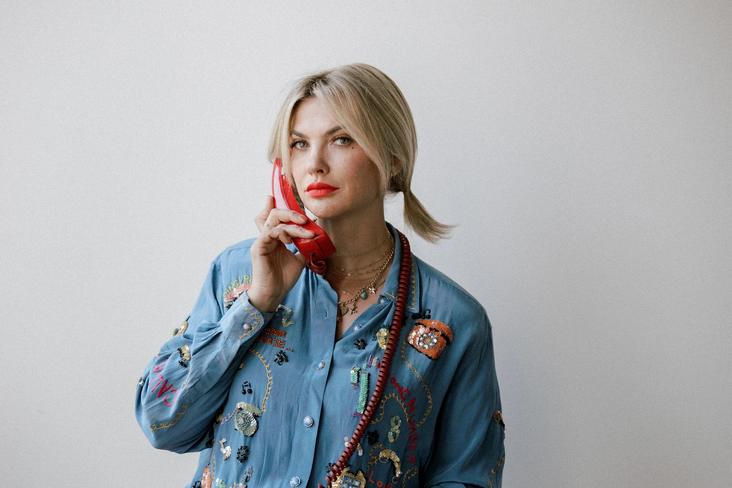 Mindy Gledhill, an indie singer-songwriter, with blonde hair in a side ponytail is holding a red rotary phone to her ear, dressed in a blue embroidered shirt with various patches and jewelry, against a plain gray background.