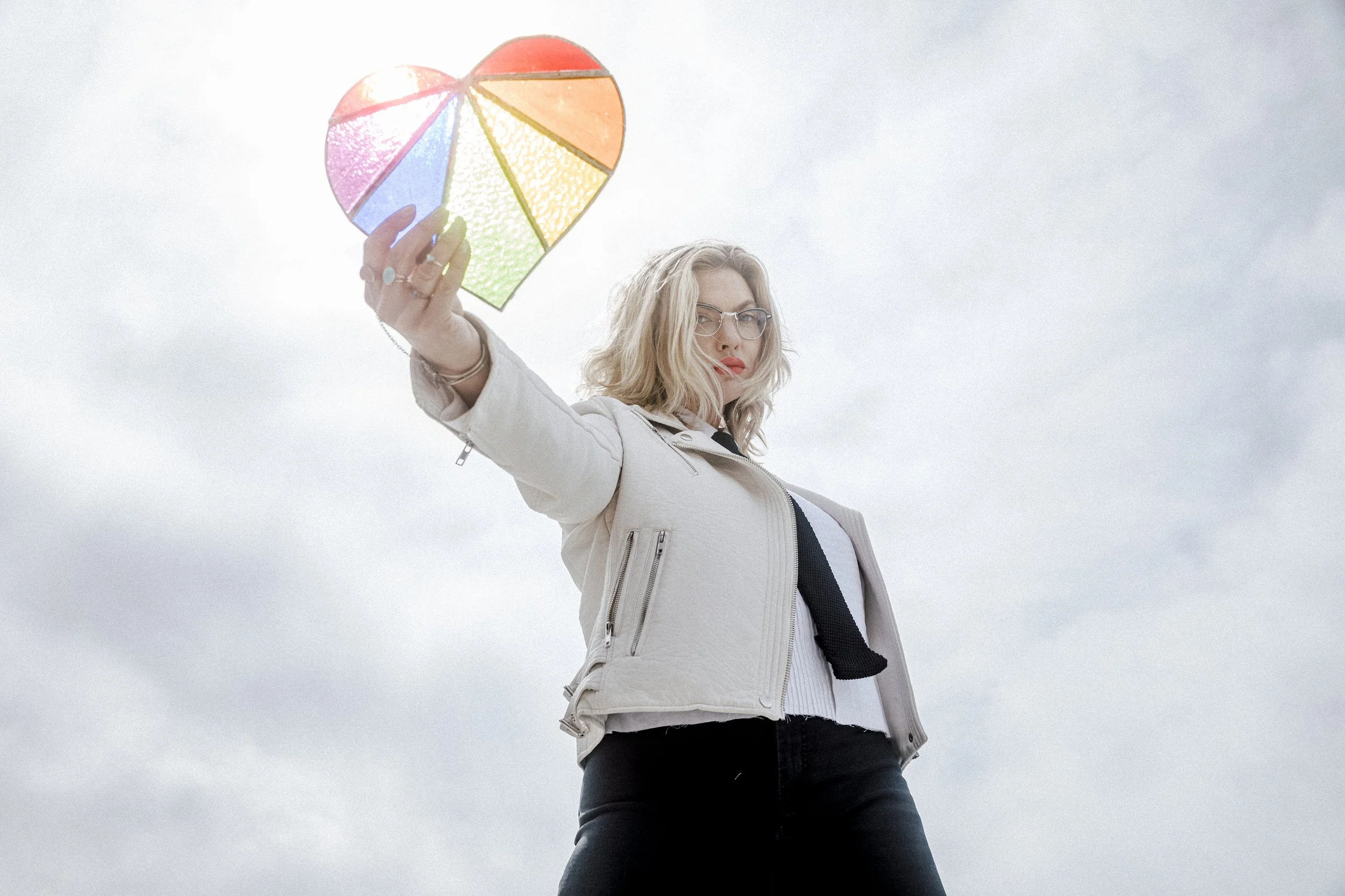 Mindy Gledhill, an indie singer-songwriter, with blonde hair and glasses holding a colorful heart-shaped lantern outdoors against a cloudy sky.