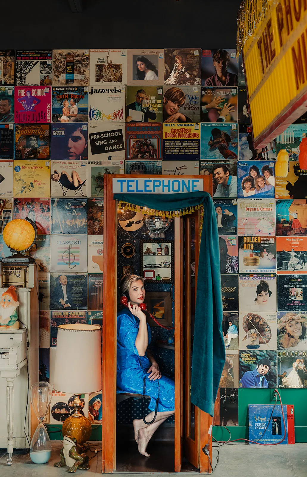 A young woman dressed in a blue dress sitting inside a vintage phone booth, holding a red telephone receiver to her ear, with a wall decorated with vintage album covers in the background.