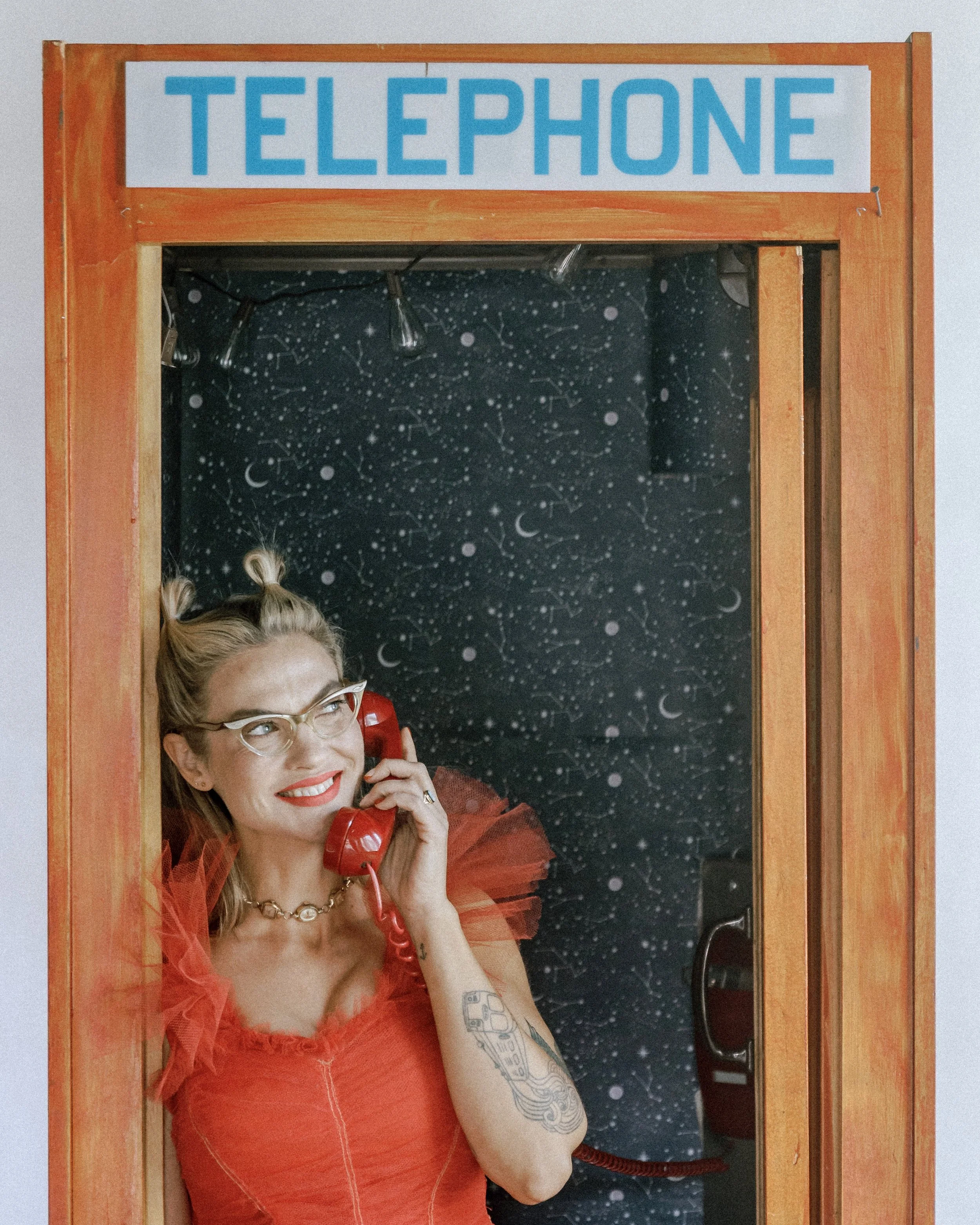 Mindy Gledhill, an indie singer-songwriter,  with glasses and tattoos wearing a red dress, talking on a red pay phone inside a vintage phone booth with a galaxy-themed background.
