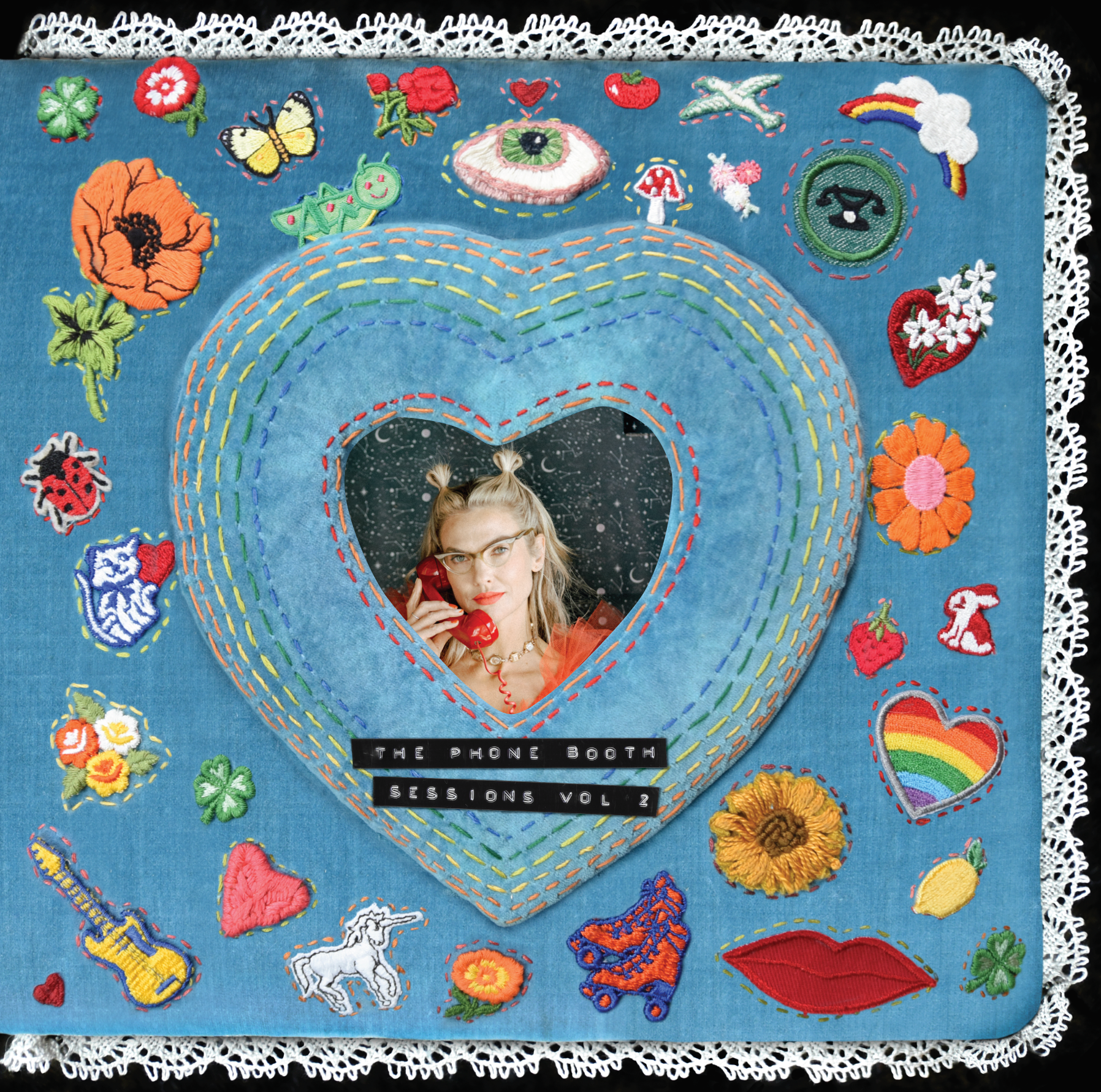 A fabric-covered book with embroidered colorful patches and a heart-shaped mirror. The mirror reflects Mindy Gledhill holding a red telephone. The book has a lace trim and text that reads 'The Phone Booth Sessions Vol 2'.