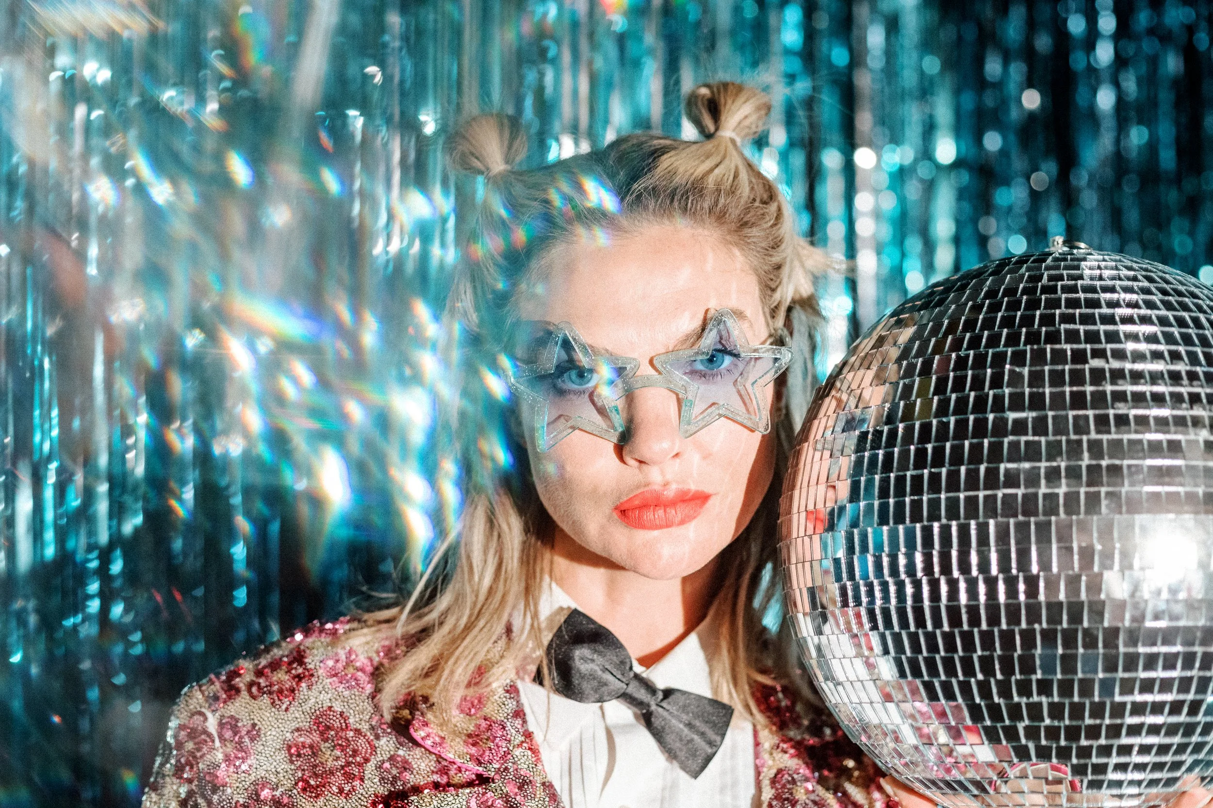 Mindy Gledhill, an indie singer-songwriter, in star-shaped sunglasses holds a large disco ball, with a shiny, blue tinsel curtain in the background. She wears a glittery blazer, a white shirt, a black bowtie, and has her hair styled in space buns.