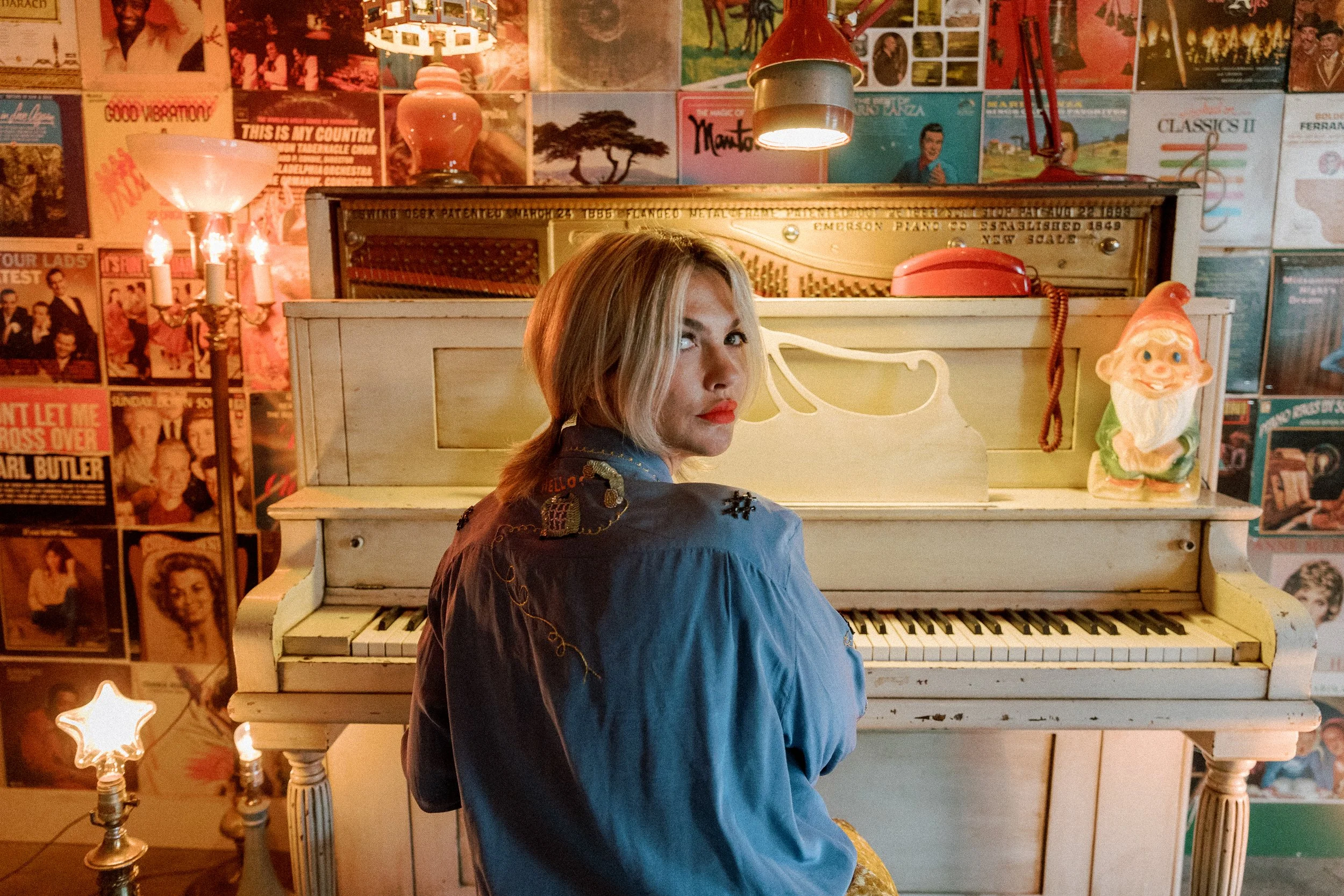 Mindy Gledhill with blonde hair seated at a vintage white piano, looking over her shoulder. The room is decorated with colorful posters and album covers on the wall, with warm lighting including a red desk lamp and a small star-shaped lamp.