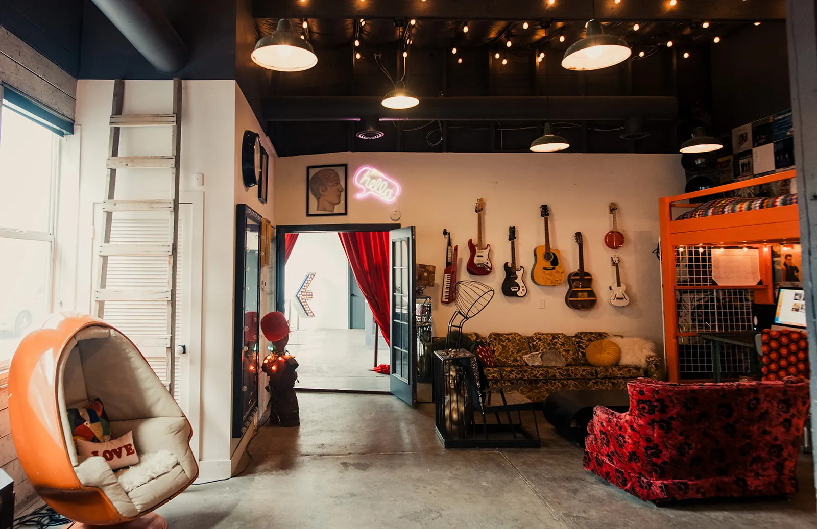 Interior of a colorful music-themed room featuring a wall with multiple guitars, a vintage sofa, vibrant patterned chair, decorative pillows, wall art including a framed face and a neon sign saying 'hello'.