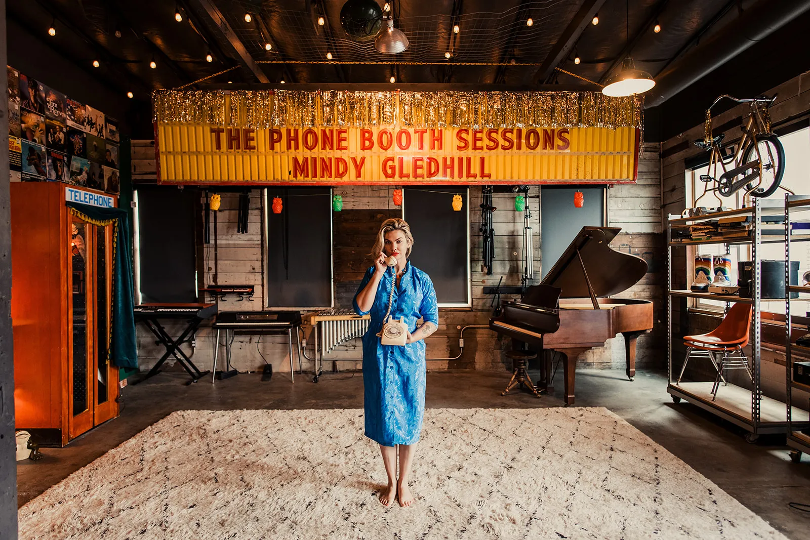 A woman in a blue dress holding a vintage rotary phone, standing in a room with musical instruments and a colorful sign that reads 'The Phone Booth Sessions, Mindy Gledhill'.