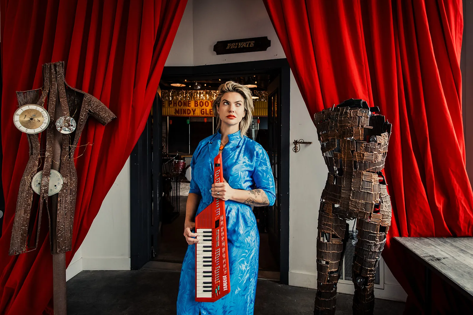 A woman standing in front of a doorway with a sign that reads 'PHONE BOOTH MINDY GLE.' She is holding a red Yamaha keyboard prop and is wearing a bright blue dress. The background features red curtains, a vintage dress form, and decorative art pieces