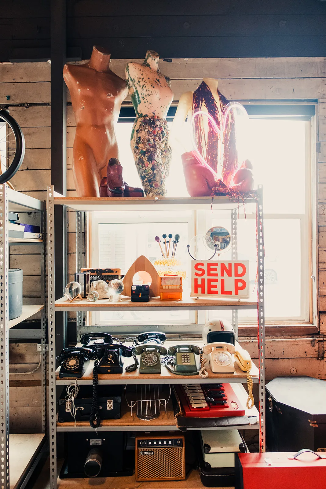 Shelves with vintage phones, a sign that says 'SEND HELP,' mannequin torsos, shoes, neon heart sign, and decorative objects in a thrift store.