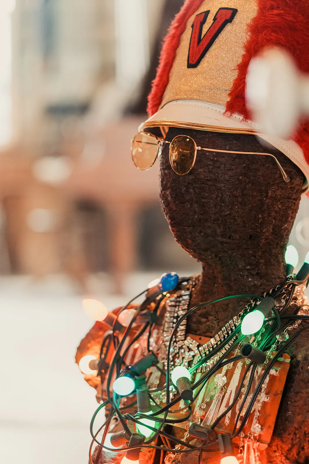 A mannequin head wearing a red and white athletic cap with a red letter V, round sunglasses, multiple strands of Christmas lights, and layered necklaces.