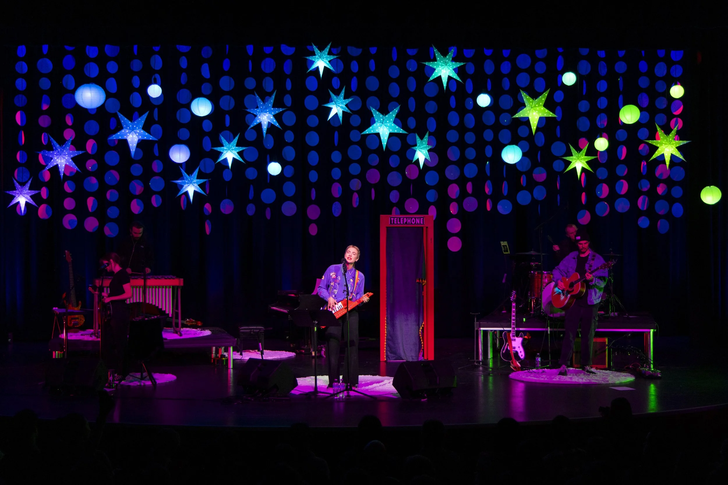 A band performing on stage with colorful star and circle decorations hanging above, including a woman singing and playing a keytar, and a man playing an acoustic guitar, with musical instruments and equipment around them.