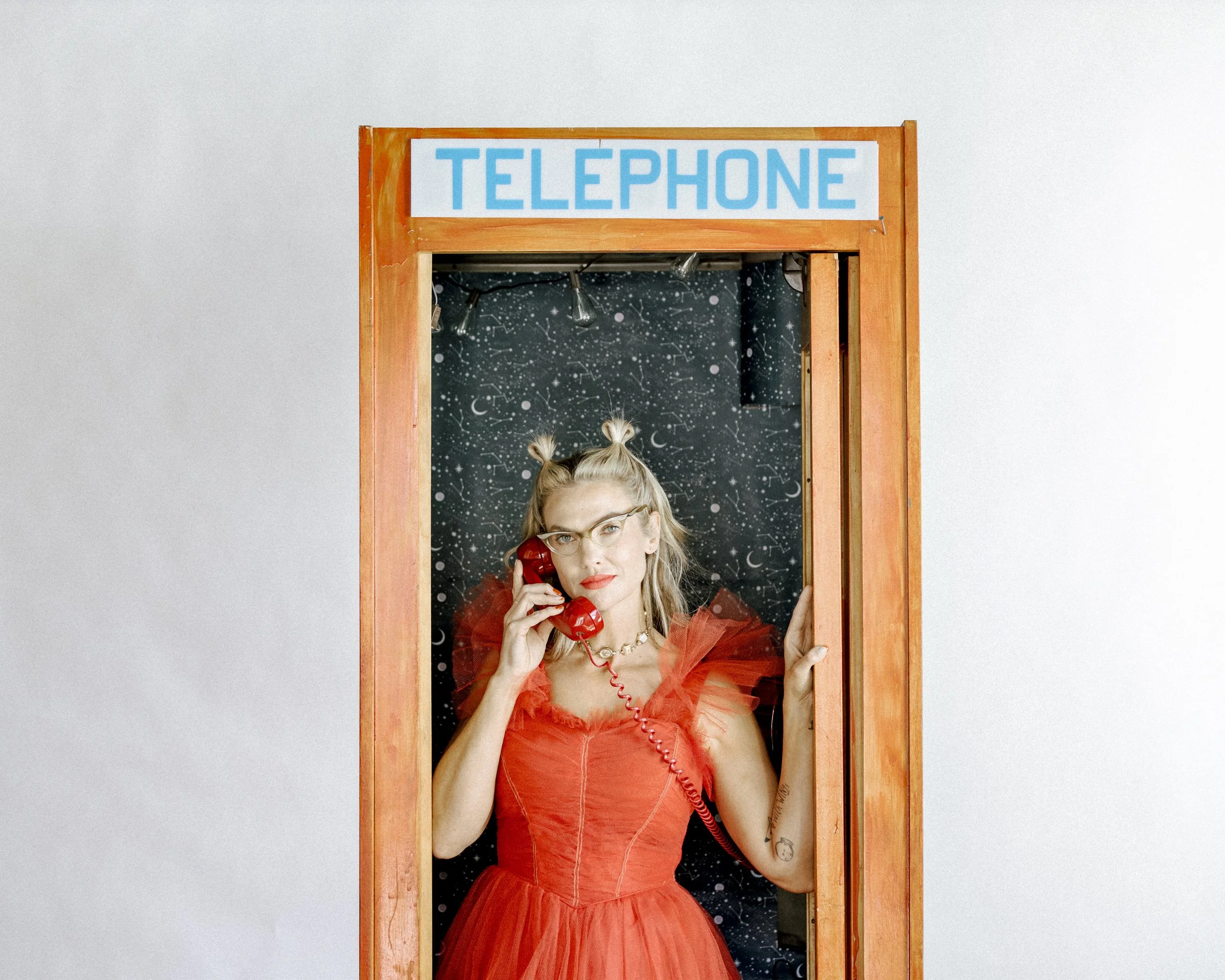 Mindy Gledhill, an indie singer-songwriter, with blonde hair styled in space buns, wearing glasses and a red dress, talking on a red telephone inside a small wooden phone booth with a starry sky pattern background.