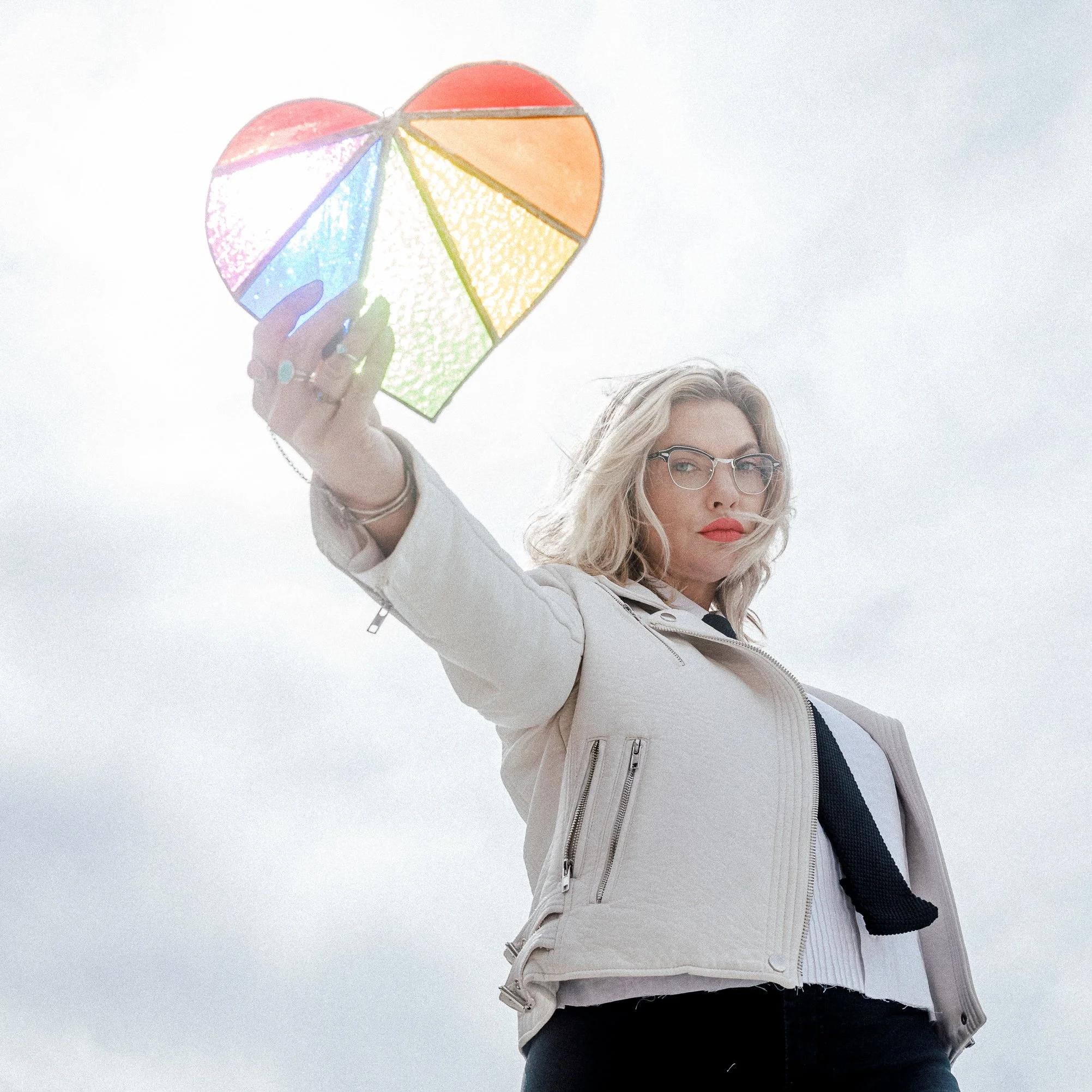 Mindy Gledhill, an indie singer-songwriter, with blonde hair, glasses, and red lipstick, wearing a beige jacket and black pants, holds a colorful, stained glass heart-shaped object up against a cloudy sky.