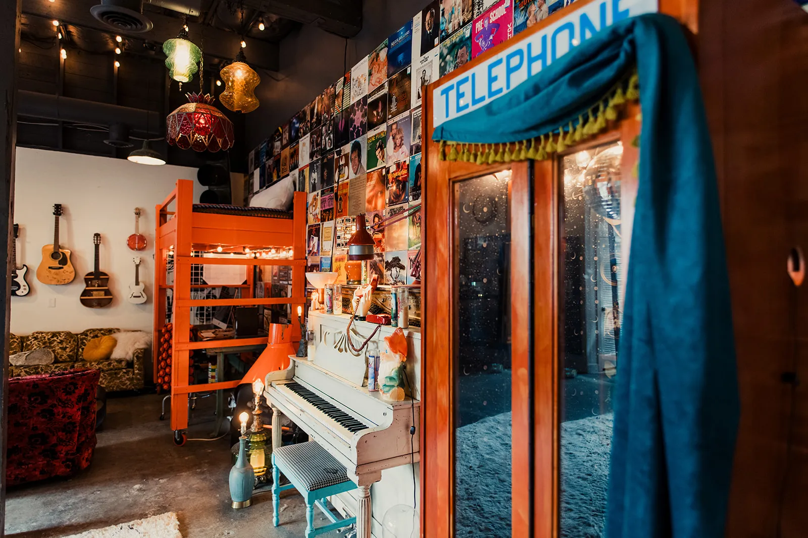 Colorful and eclectic interior room with musical instruments, including guitars on the wall, a white piano decorated with a unicorn head, and a rack of hanging lamps with fabric coverings. The wall is decorated with numerous album covers and posters,