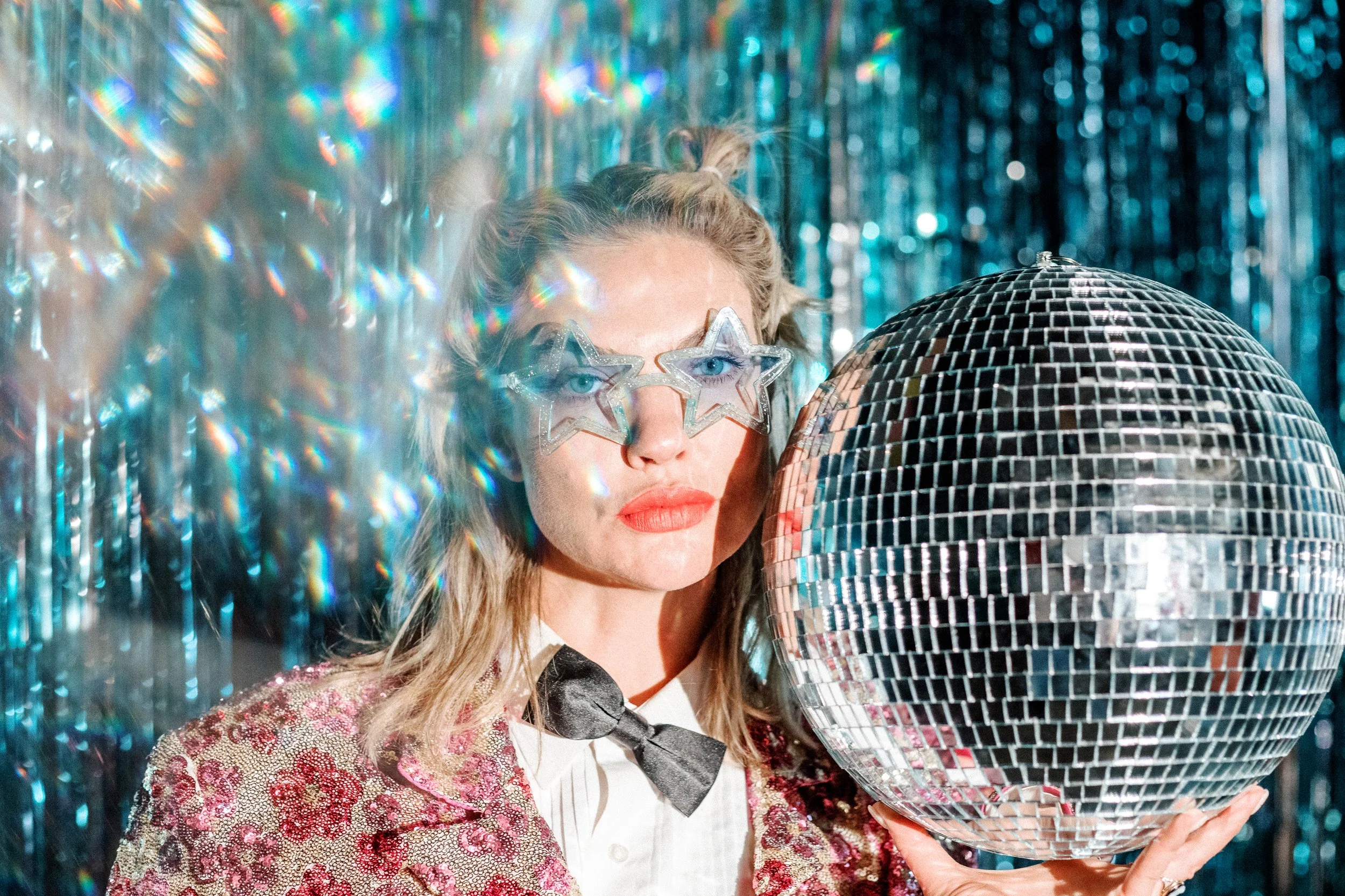 Mindy Gledhill, an indie singer-songwriter, with star-shaped glasses and bright red lipstick holds a large disco ball in front of a shiny, metallic backdrop.