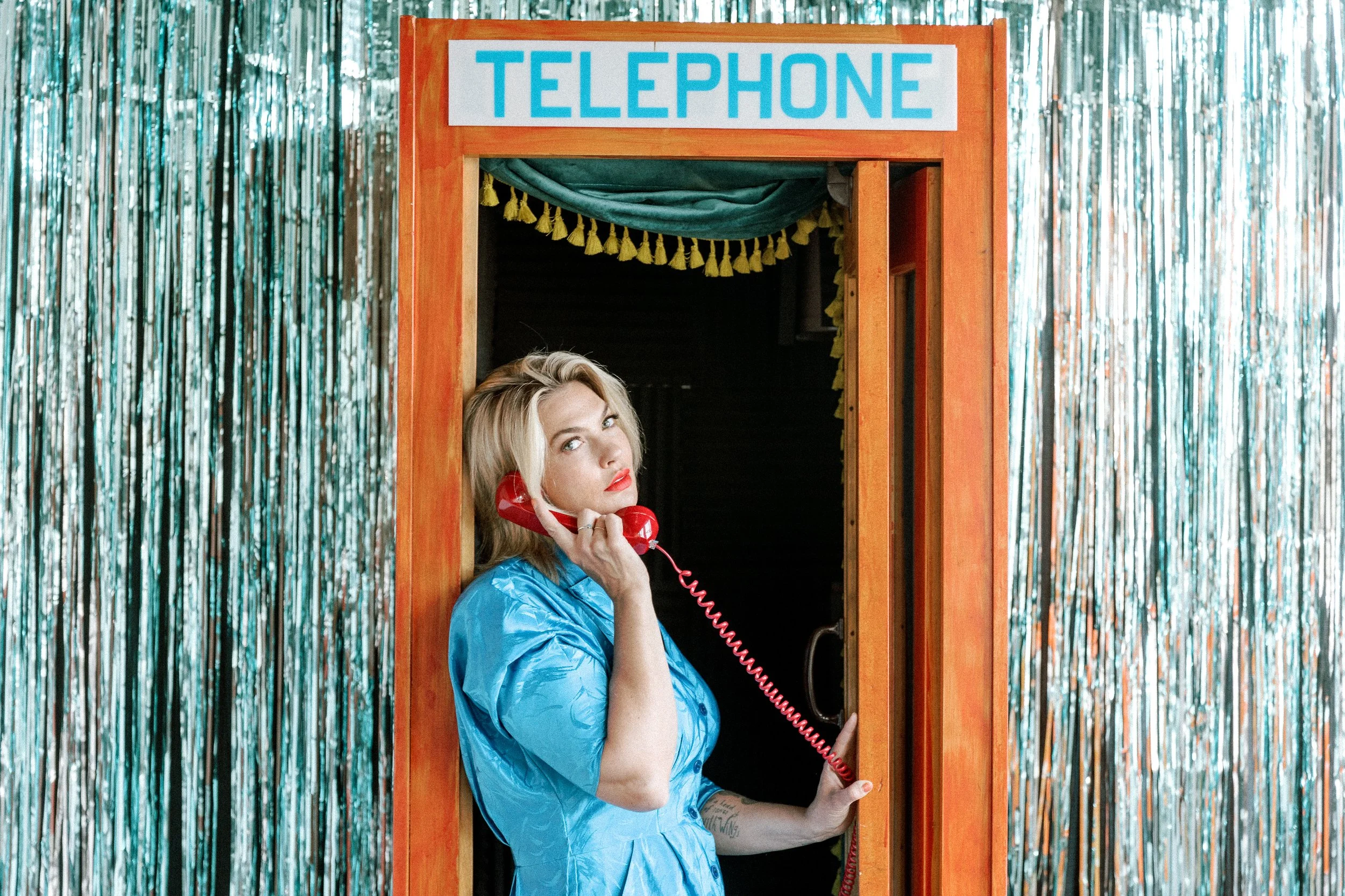 Mindy Gledhill with blonde hair and red lipstick wearing a blue dress, speaking on a red telephone inside a vintage wooden telephone booth with a blue and white "TELEPHONE" sign, against a background of silver metallic streamers.