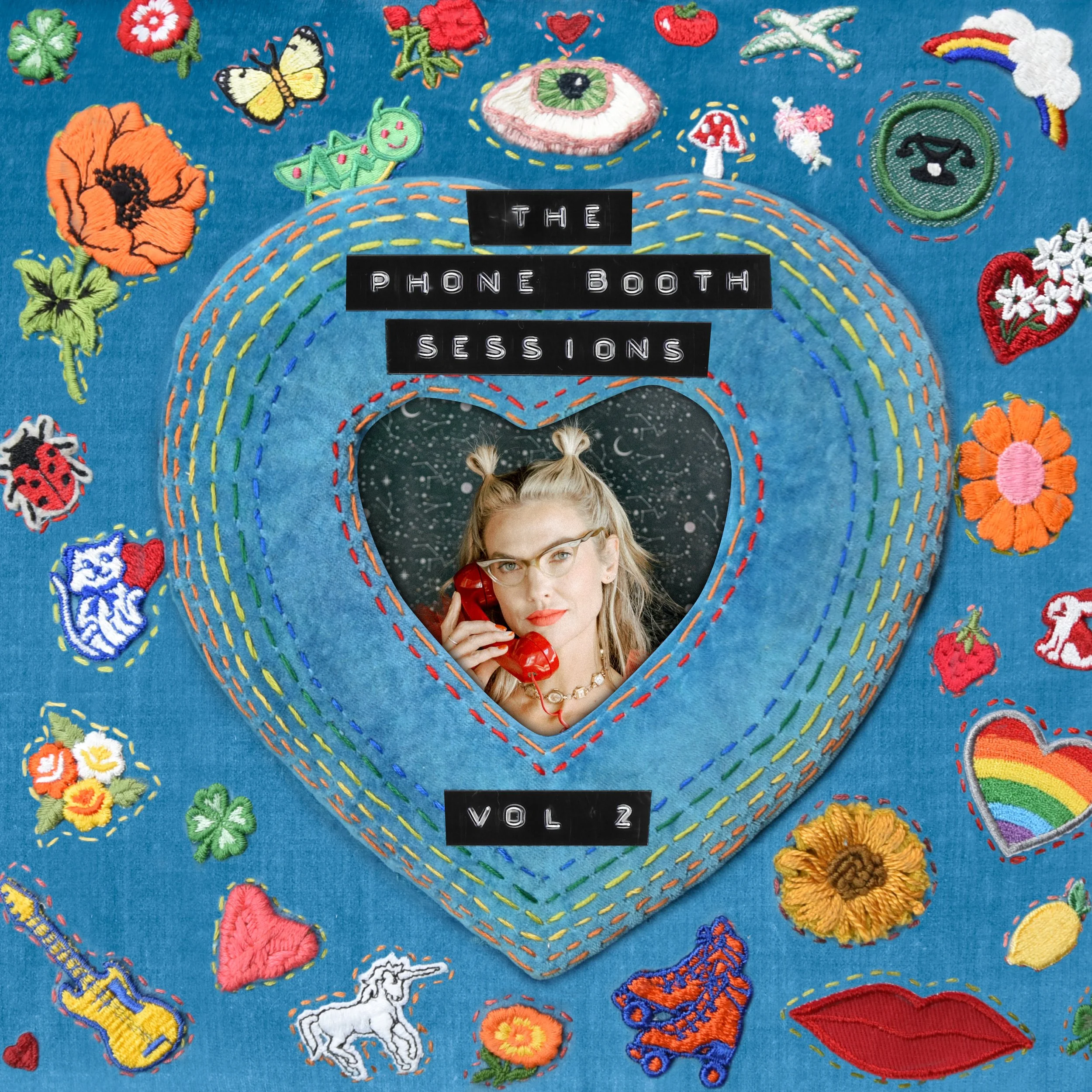 A woman with glasses and blonde hair styled in two buns, holding a red rotary phone, is framed within a heart-shaped embroidery on a blue fabric background. The embroidery is decorated with various colorful patches like flowers, a rainbow, animals, and other whimsical designs. Text above and below the woman reads 'THE PHONE BOOTH SESSIONS VOL. 2'.