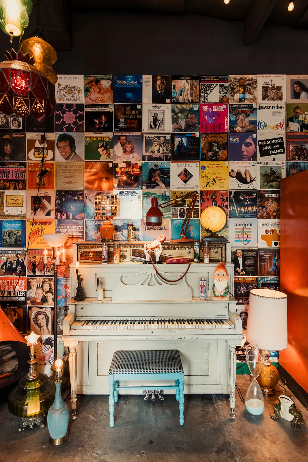 An eclectic room featuring a vintage white piano, surrounded by various lamps, vintage decor, and a wall covered with numerous colorful records and album covers.