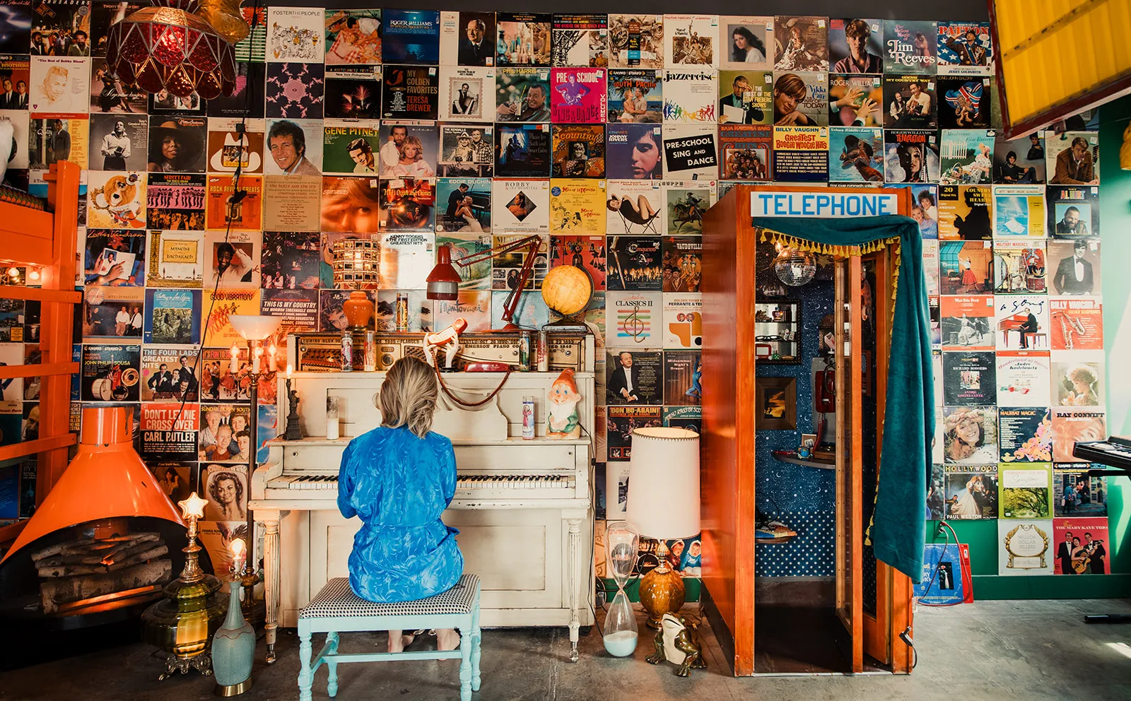 A person with blonde hair sits at a white piano with their back to the camera, wearing a bright blue dress. The room is decorated with a wall covered in colorful vintage record album covers, and various eclectic items like a globe, lamps, and a telep