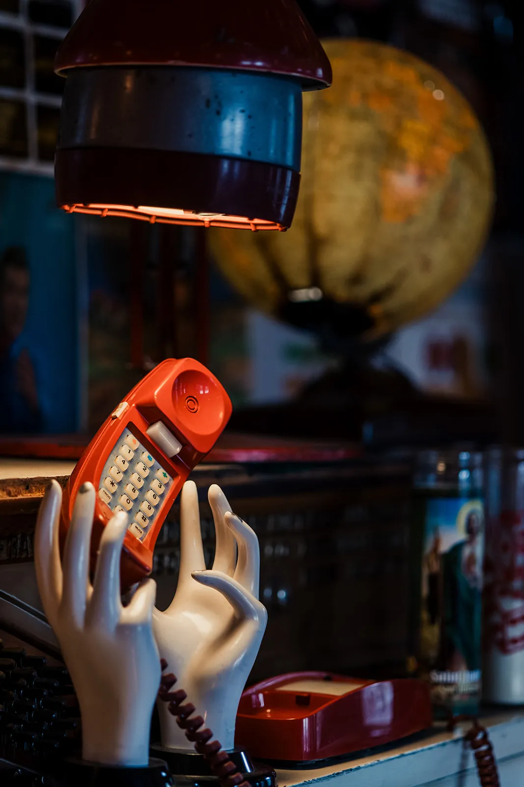 A vintage orange telephone with a rotary dial held by a white ceramic hand model, placed on a shelf with a large globe and other objects behind it.