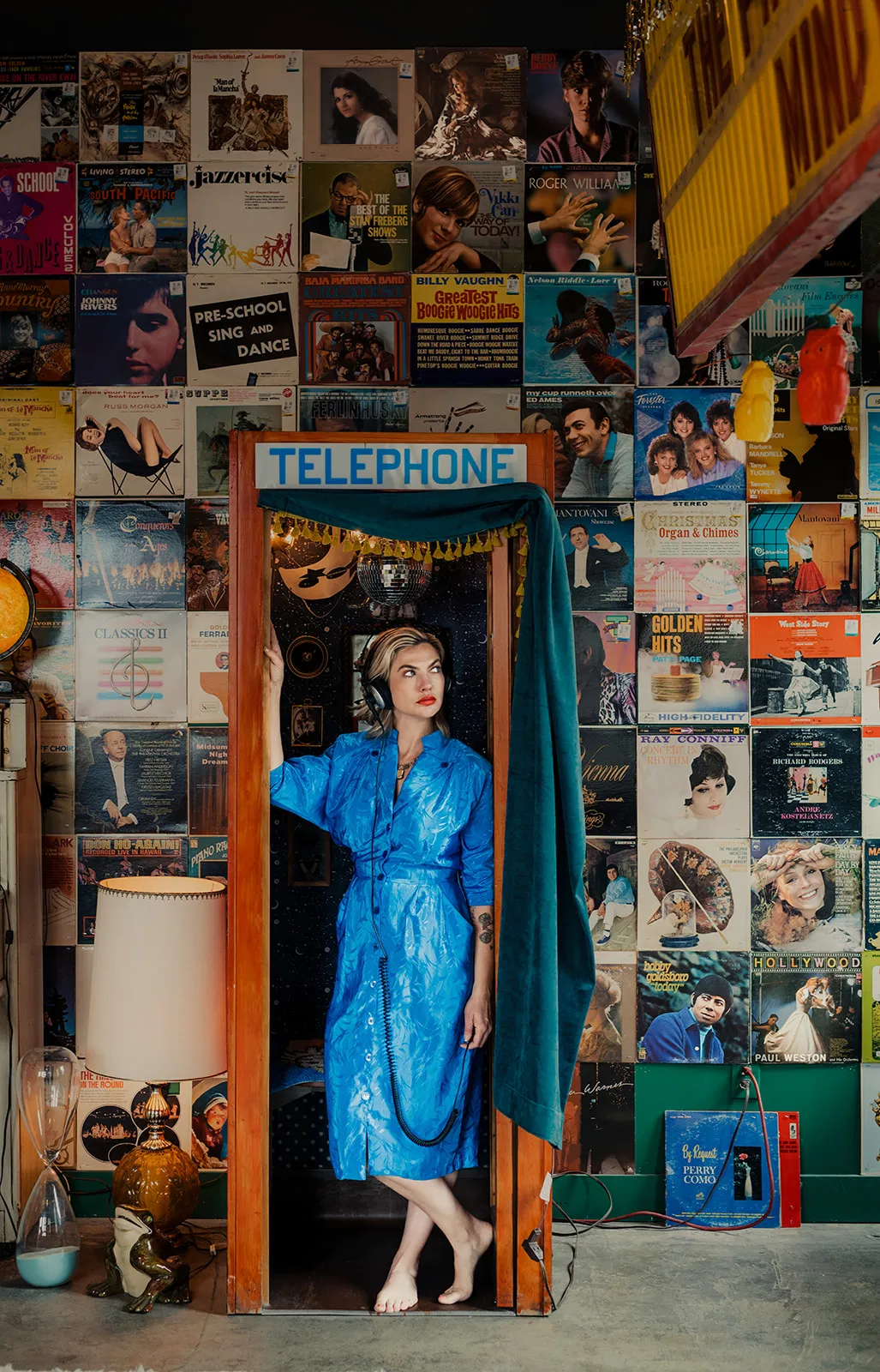 A woman with blonde hair wearing a blue dress and headphones stands inside a vintage telephone booth labeled 'TELEPHONE' in a room decorated with a wall full of vintage vinyl records and album covers.